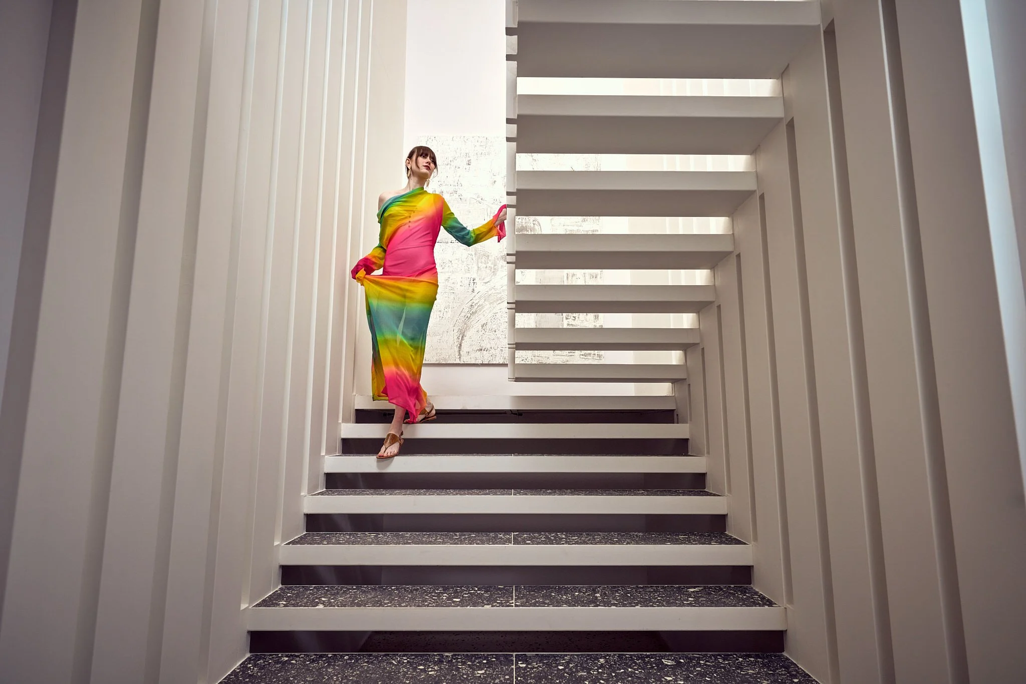 A woman in a colorful rainbow dress standing on a staircase in a modern indoor setting.