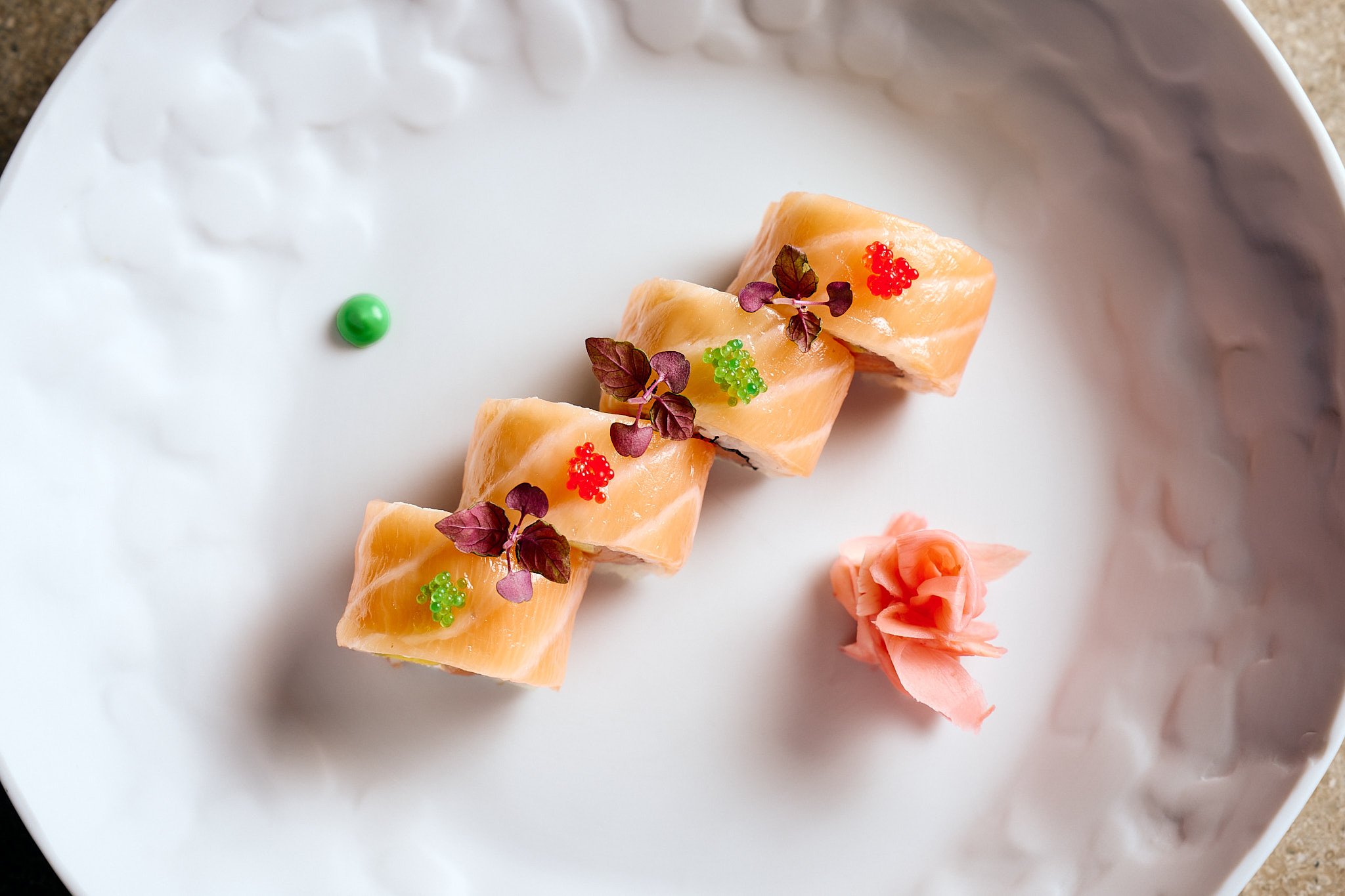 Slices of salmon topped with microgreens and colorful fish roe, served with pink pickled ginger on a white textured plate.