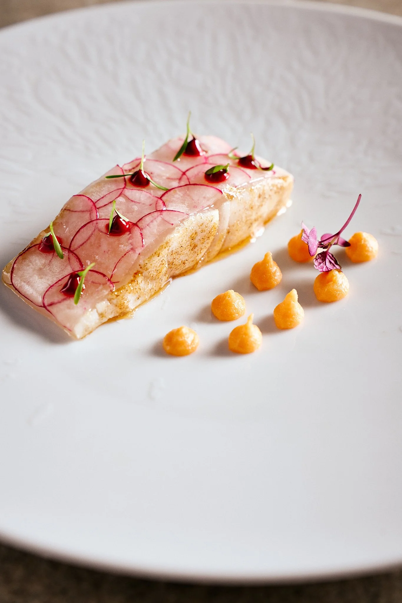Elegant plated dessert with a rectangular layer of pink and white glaze, topped with microgreens and small red garnishes, accompanied by small orange dollops and purple edible flowers on a white plate.