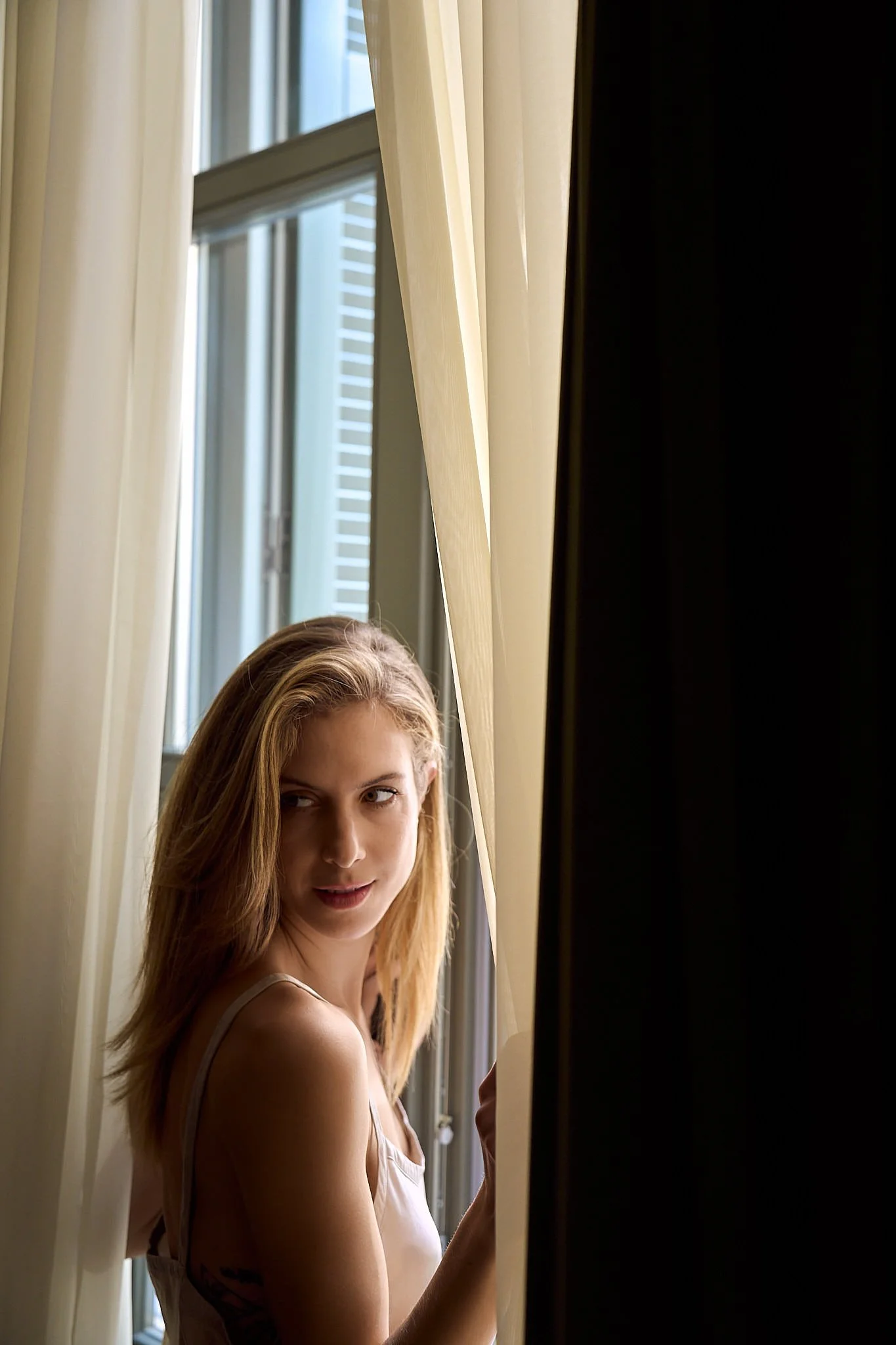 A young woman with blonde hair standing by a window with cream curtains, looking out.