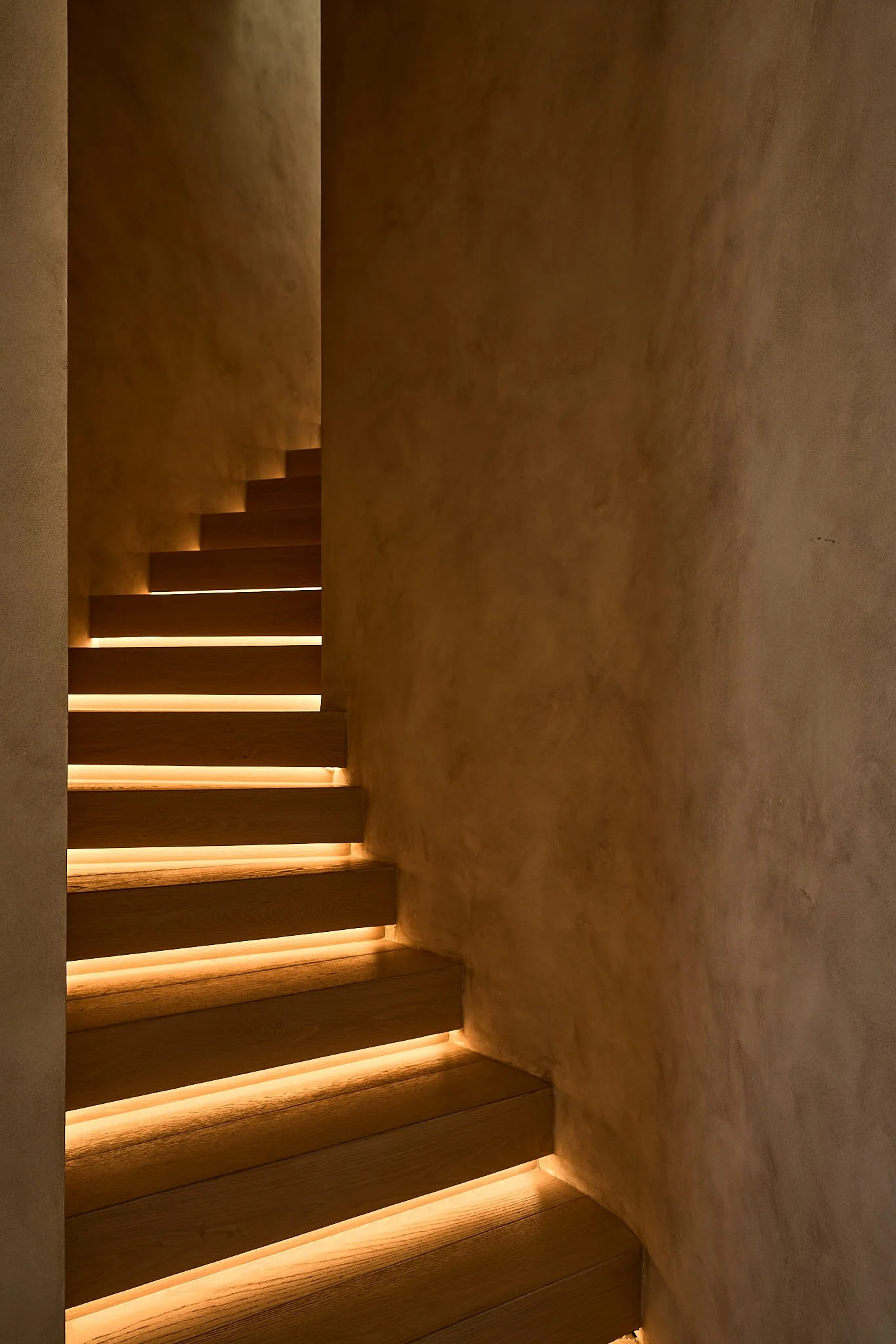 A staircase with wooden steps and soft lighting underneath each step, leading upwards along a curved wall.