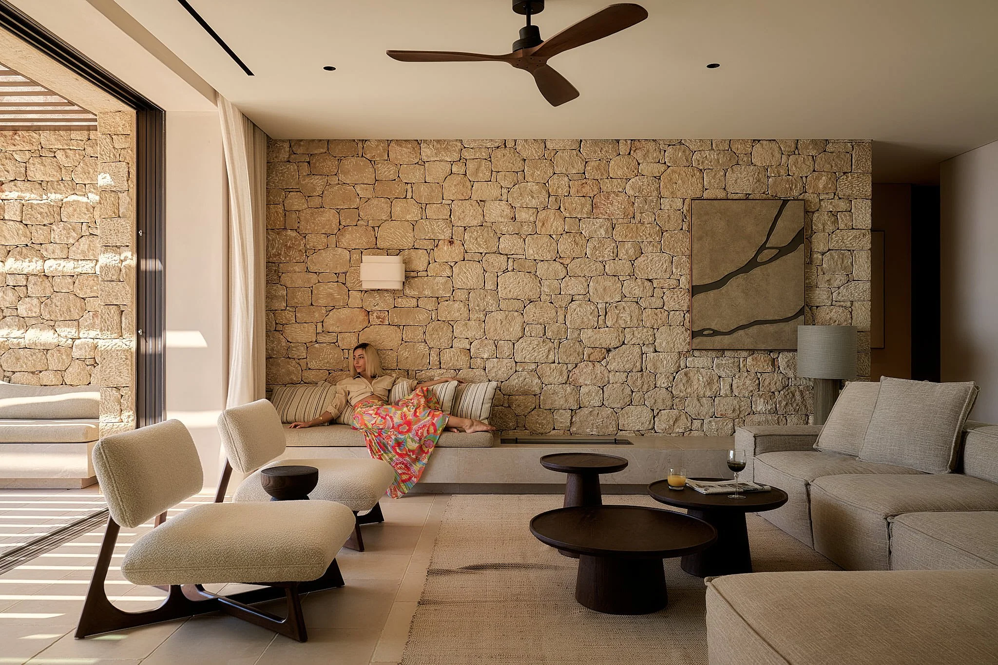 Living room with a stone accent wall, beige sofa, two beige armchairs, and a woman sitting on a built-in bench with a colorful blanket. There are three dark wooden coffee tables, a side table with a lamp, and a beige area rug. Large sliding glass doo