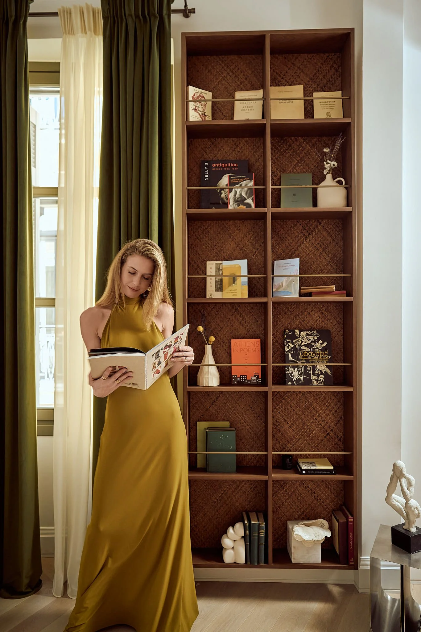 A woman in a long yellow dress reading a book or magazine while standing next to a tall wooden bookshelf filled with books and decorative items, near a window with green and white curtains.