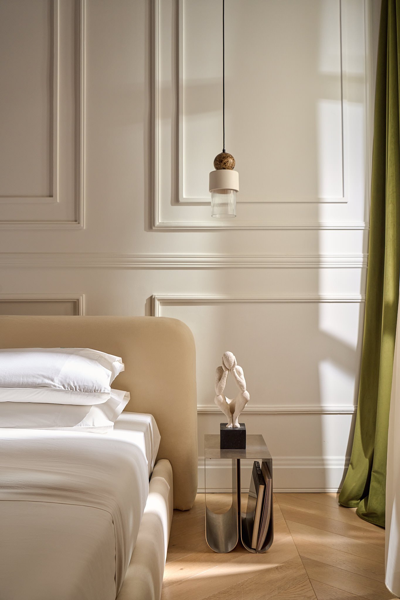 A bedroom with a beige upholstered headboard, white bedding, a small side table with a sculpture of a seated figure, and a hanging light fixture. There is a window with green curtains on the right side.