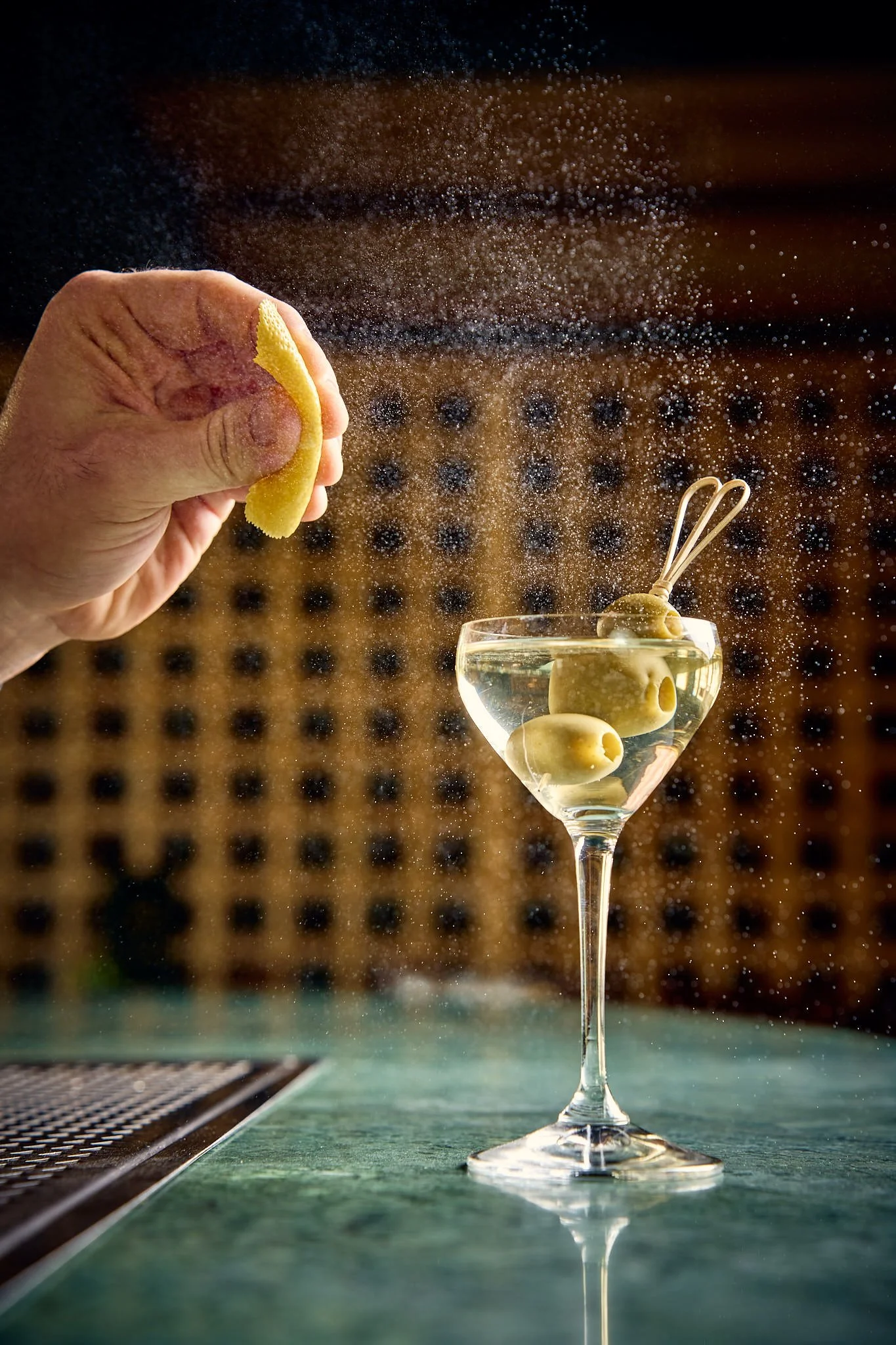 Close-up of a hand squeezing a lemon into a cocktail glass with olives, with lemon zest in the air.