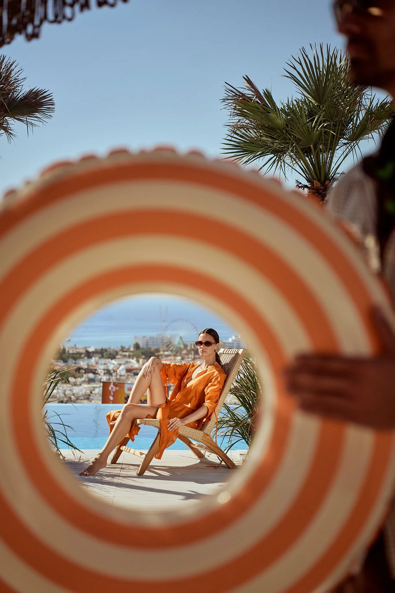 Woman lounging on a wooden chair by a pool, wearing an orange dress and sunglasses, seen through a striped orange and white circular frame, with palm trees and a cityscape in the background.