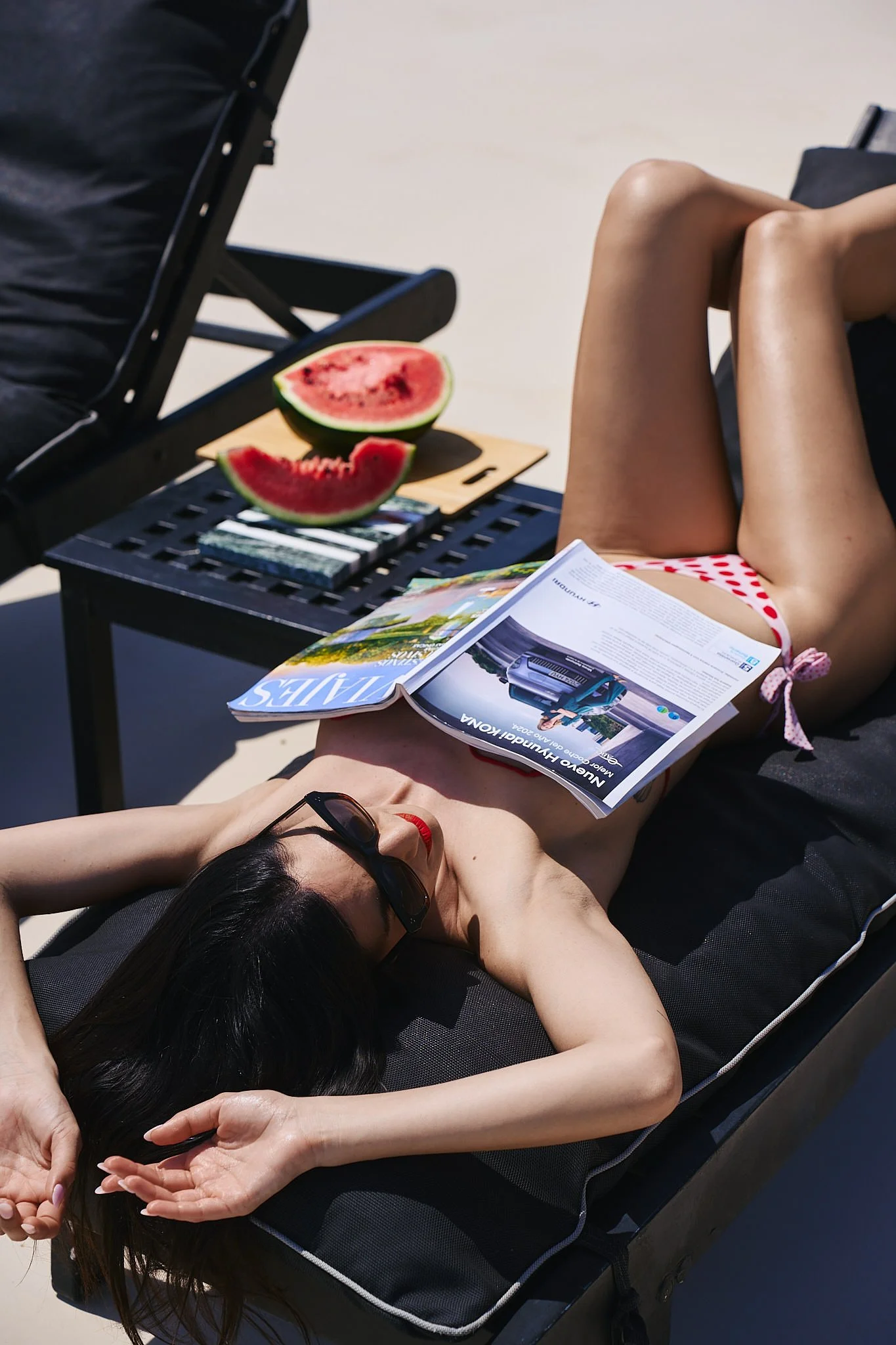 Woman sunbathing on a lounge chair at the beach, reading a magazine, with sliced watermelon on a nearby side table.