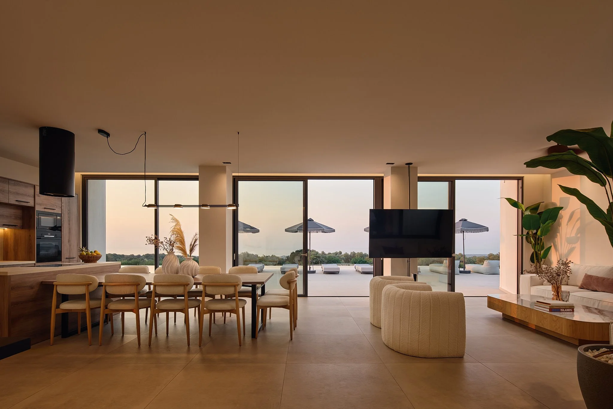 Modern living room with a dining area, large glass sliding doors opening to an outdoor patio with umbrellas, lounge chairs, and scenic view.