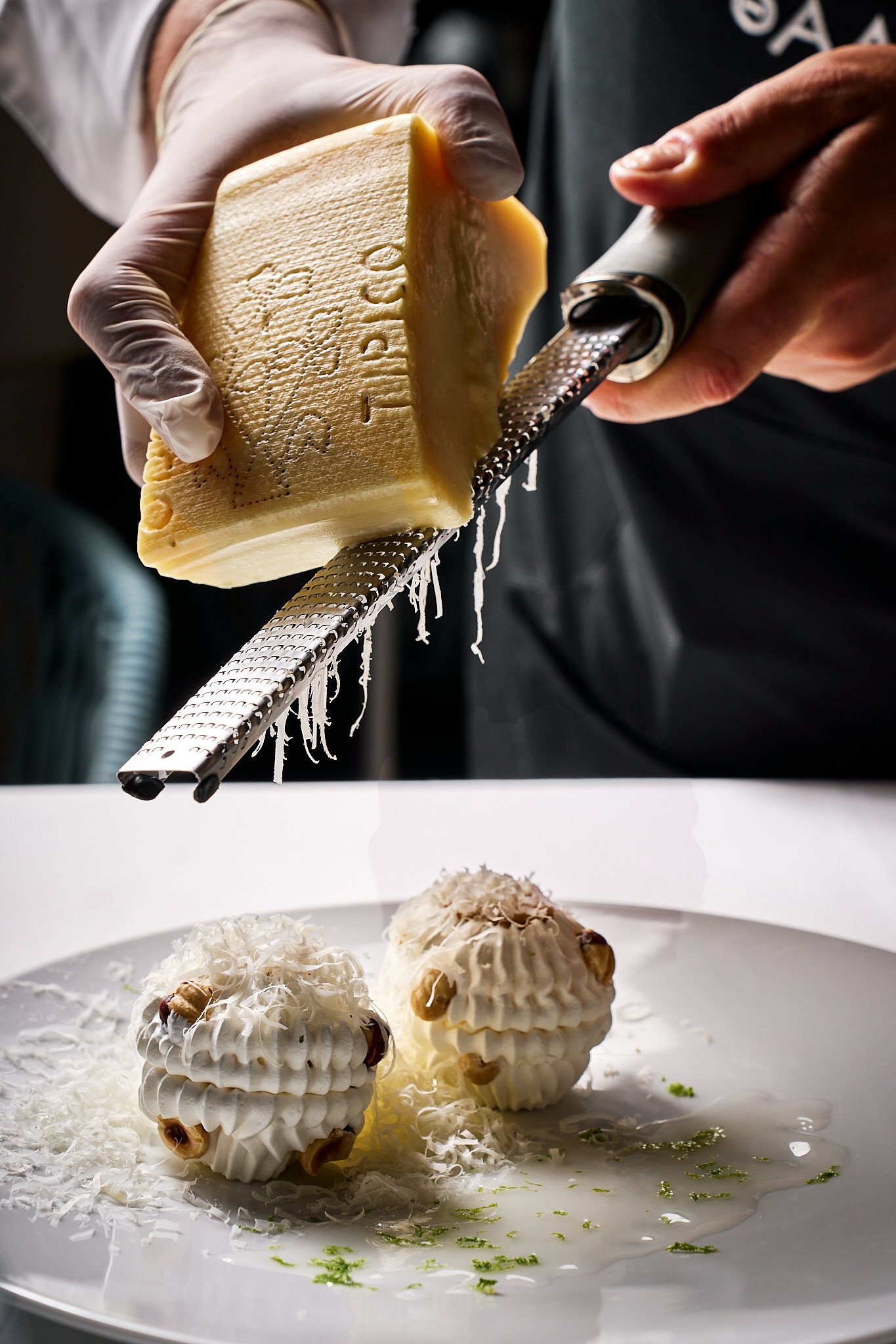 A chef grating cheese over two white whipped mousse desserts decorated with nuts on a white plate.