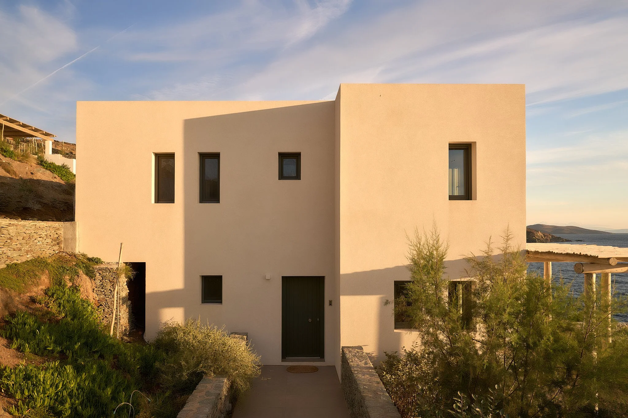 A modern white house with black window frames, situated near the water with the coastline visible in the background, during sunset.