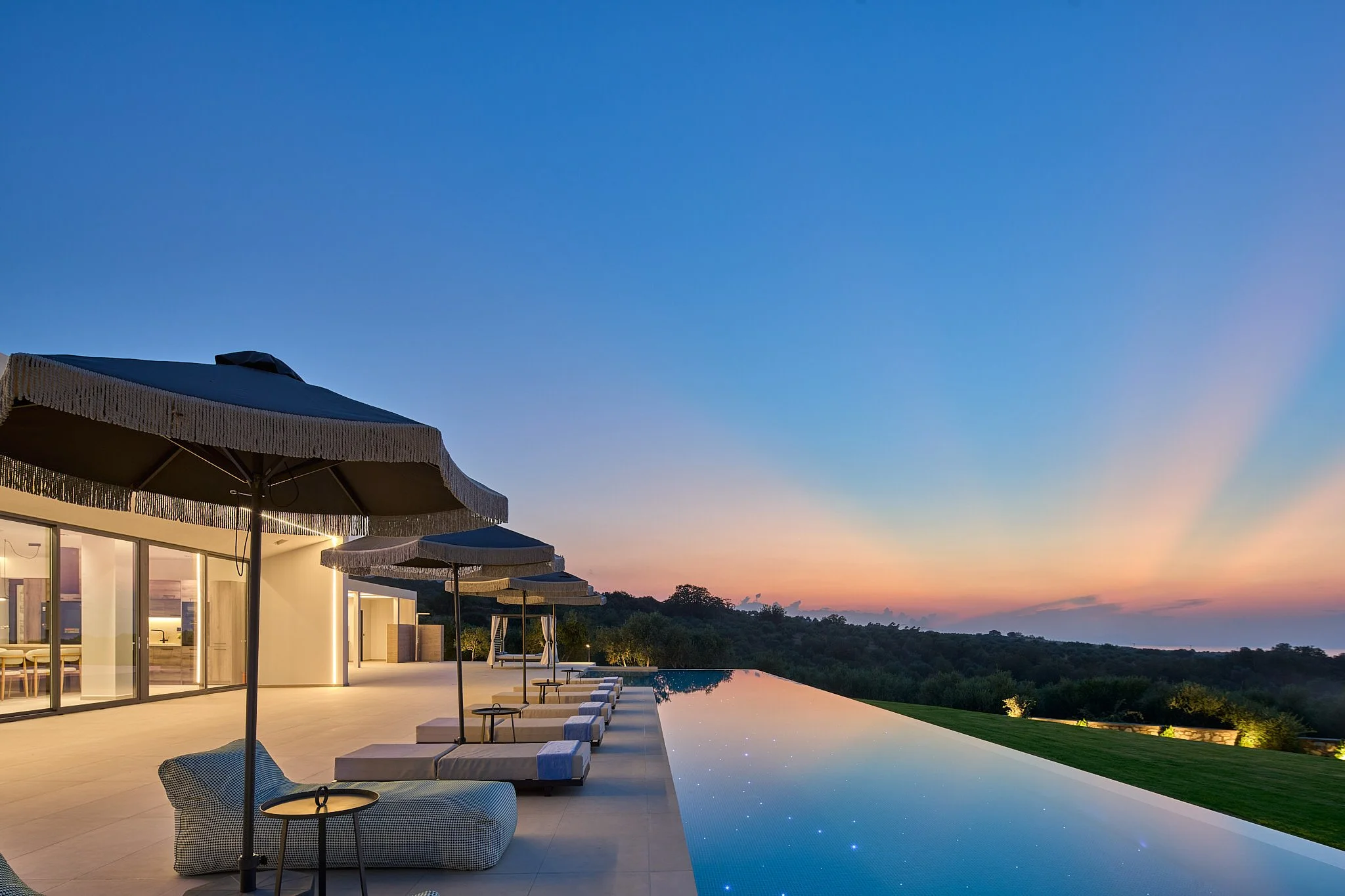 Luxury modern house with a pool overlooking a scenic landscape at sunset, outdoor lounge chairs and umbrellas on the patio.