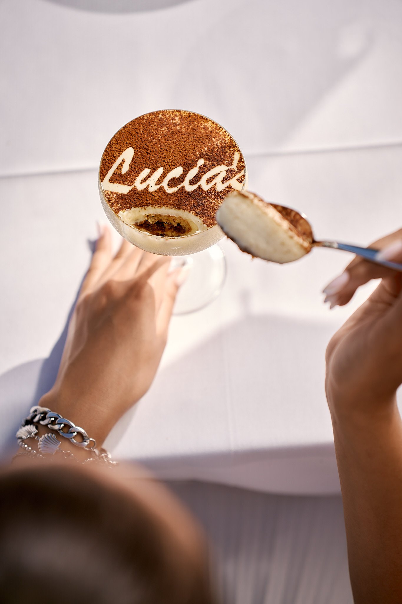 A person is holding a spoon with a piece of layered cake, and a stemmed glass with a dessert topped with cocoa powder that has the name 'Lucia' written on it, seen from above.