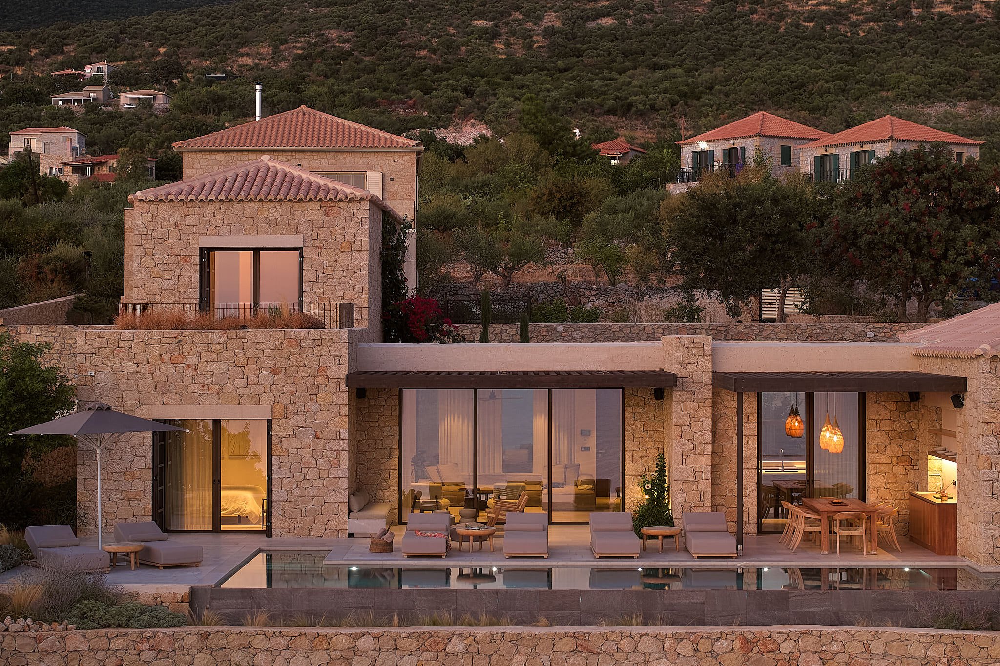 A luxurious modern house with stone walls and large glass sliding doors, featuring a patio with lounge chairs, an umbrella, a pool, and a dining area, set against a hillside with trees and other houses.