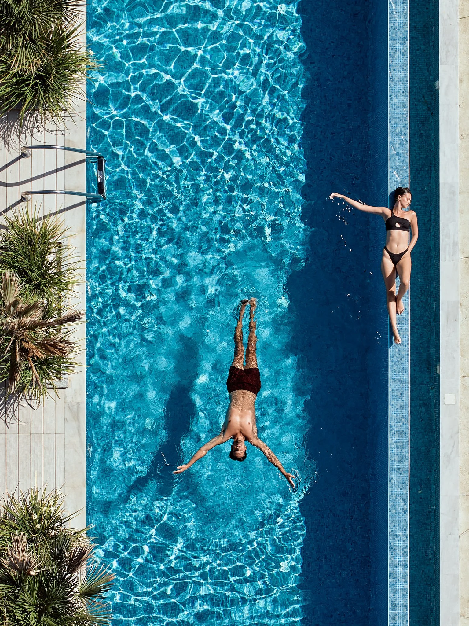 Two people relaxing by a swimming pool: a woman lying on a ledge near the pool, and a man floating on his back in the pool, surrounded by clear blue water and some greenery.