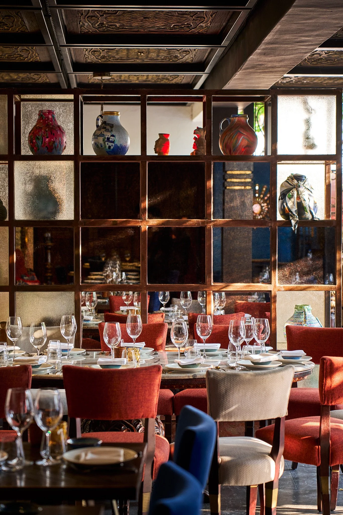 Dining area of a restaurant with wooden tables, red and beige chairs, glassware, and a decorative wooden partition with colorful vases and sculptures.