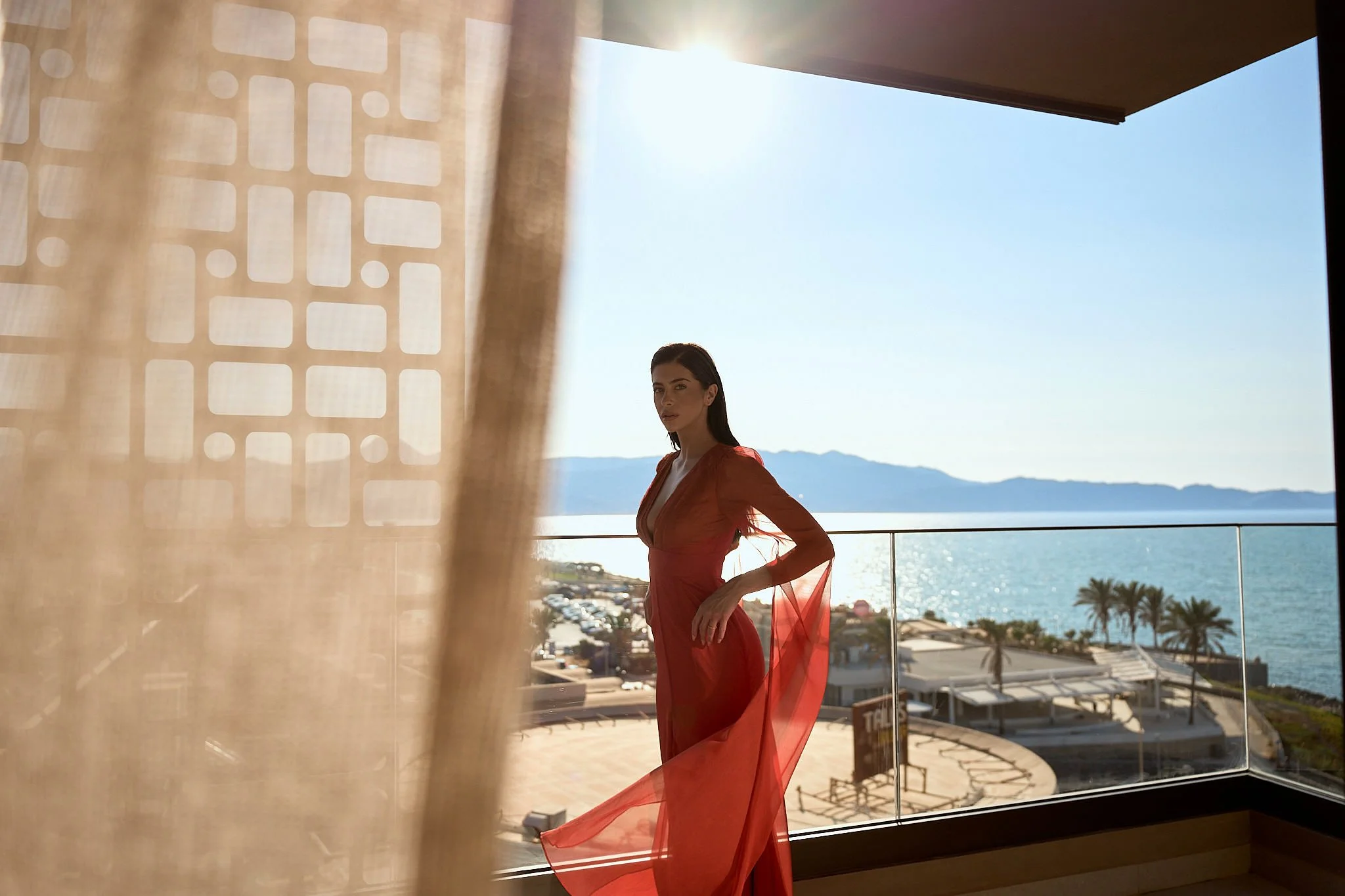 A woman in a red dress standing on a balcony overlooking the ocean and a mountainous landscape in the distance, with palm trees and a sunny sky.