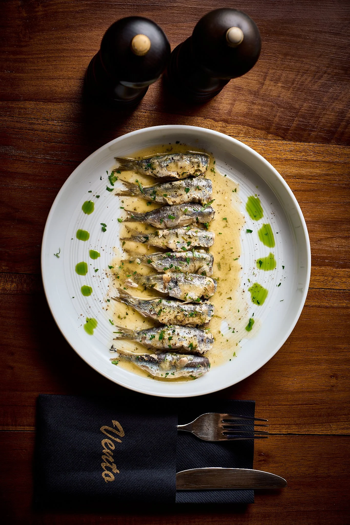 Plate of cooked sardines garnished with chopped herbs and drizzled with green oil, on a wooden table with black salt and pepper shakers in the background.