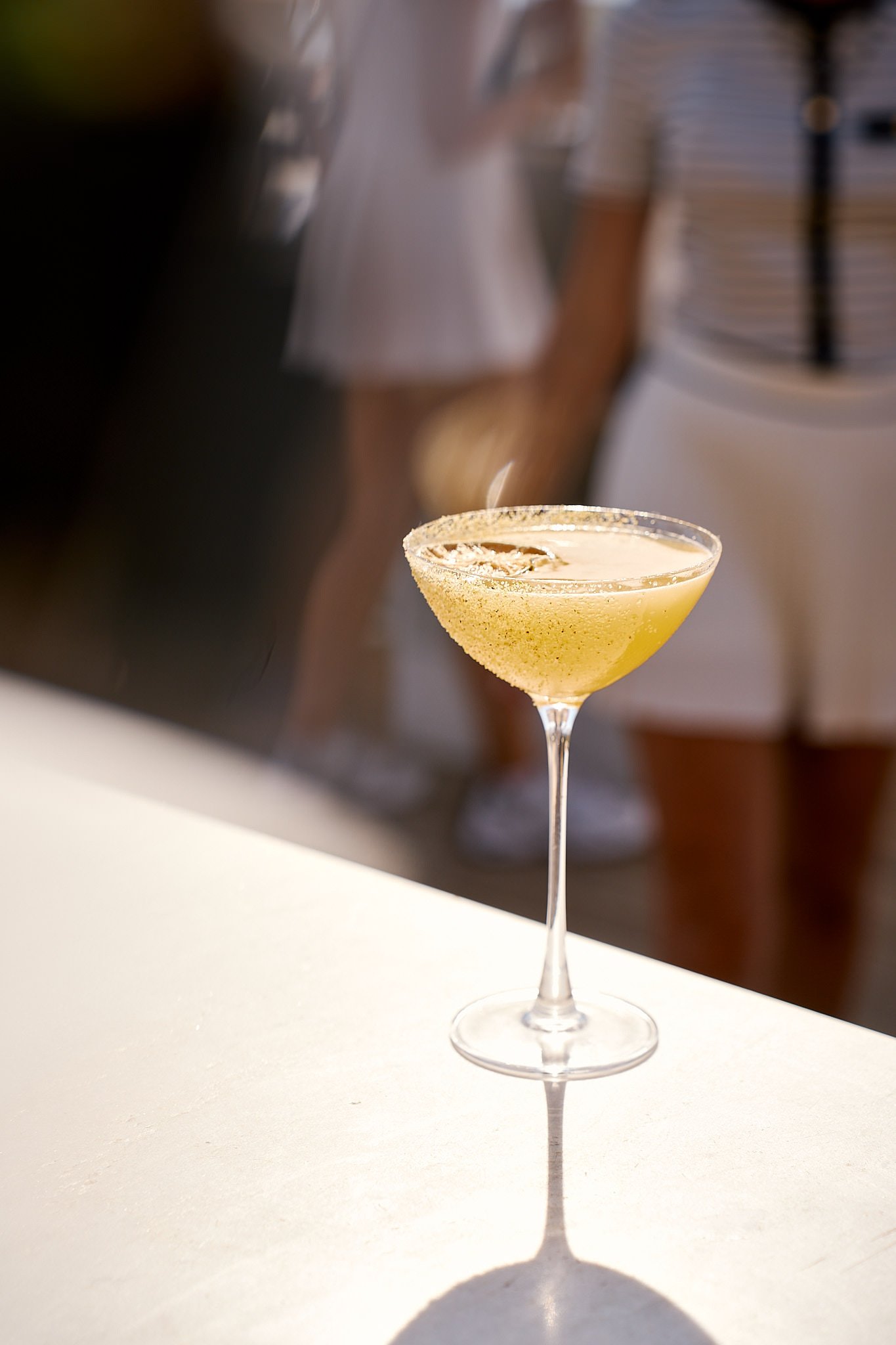 A yellow cocktail in a coupe glass with a sugar-rim and garnish, placed on a white surface with a blurred background.