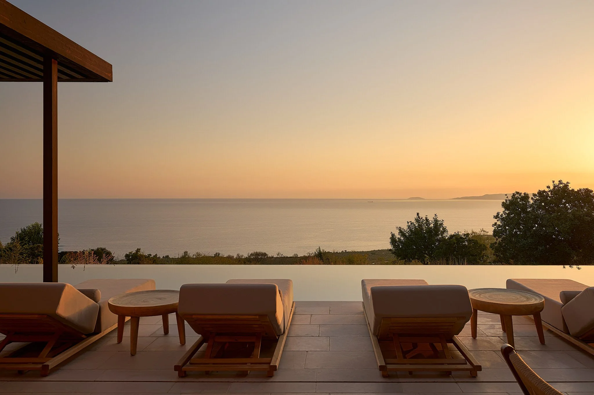 Four lounge chairs and small round tables on a deck overlooking a calm sea during sunset, with trees in the background.