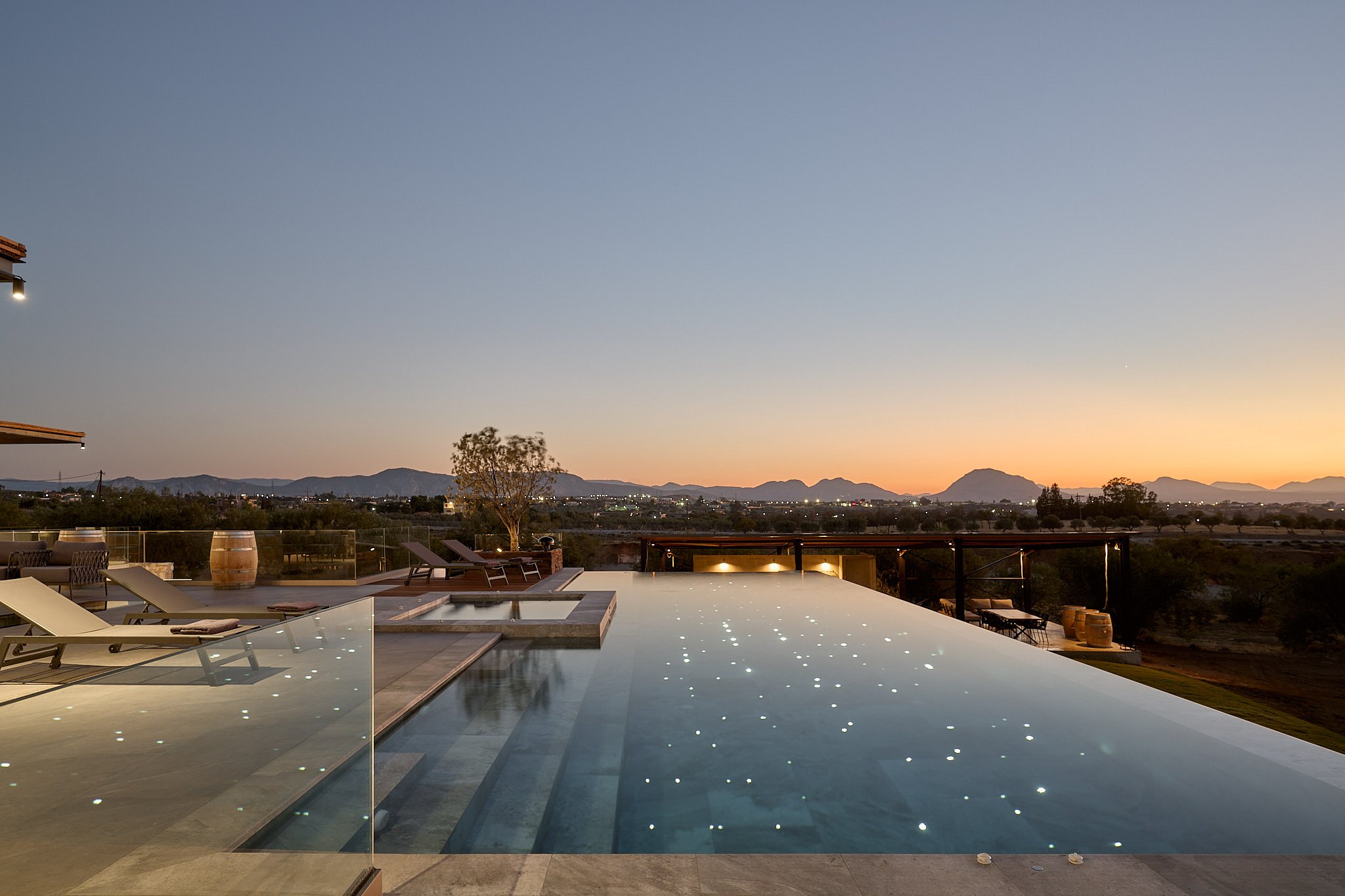 Infinity pool at sunset with lounge chairs, a tree, and distant mountains.