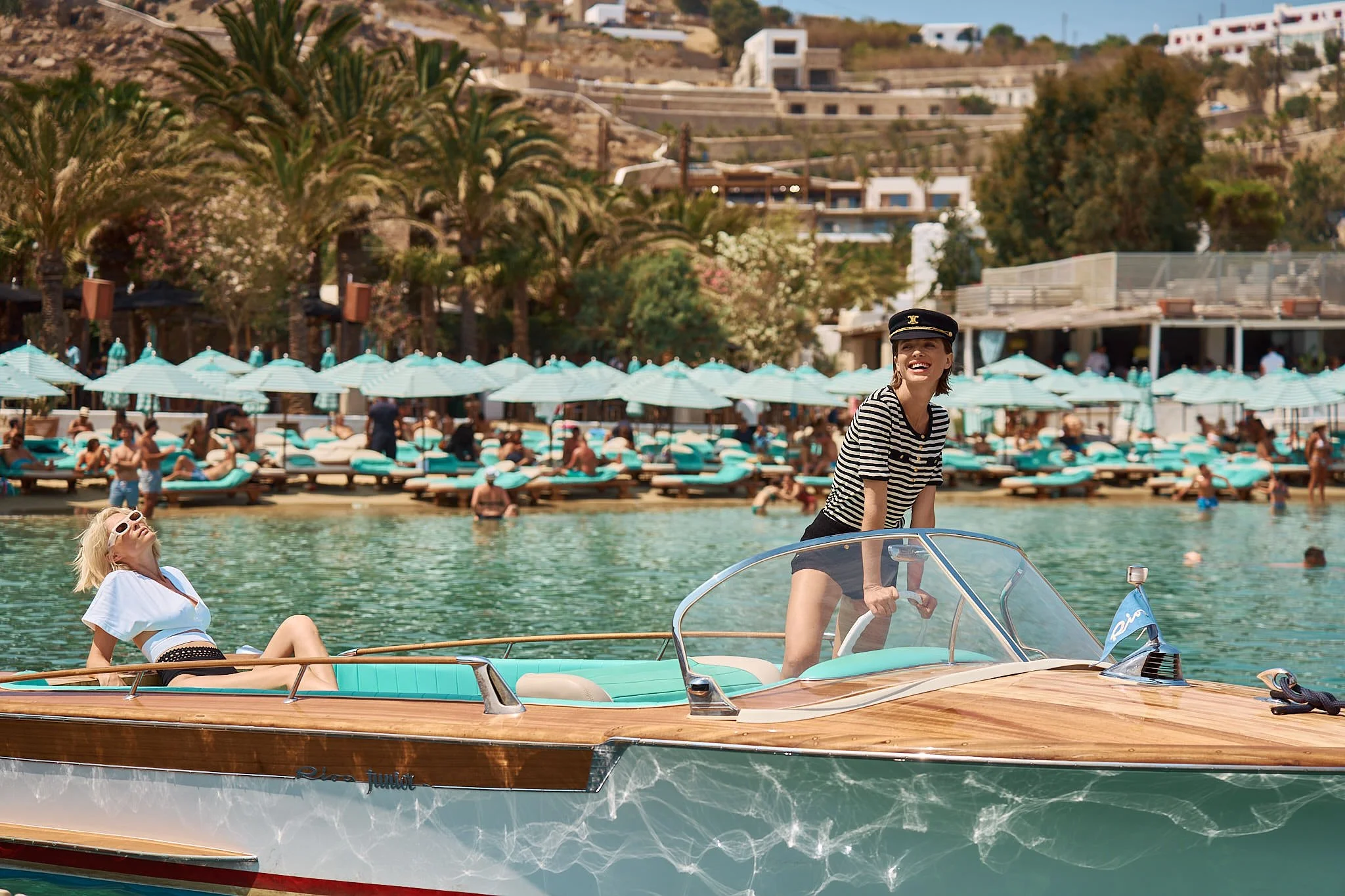 Two women on a wooden boat at a poolside resort with many lounge chairs and umbrellas in the background.