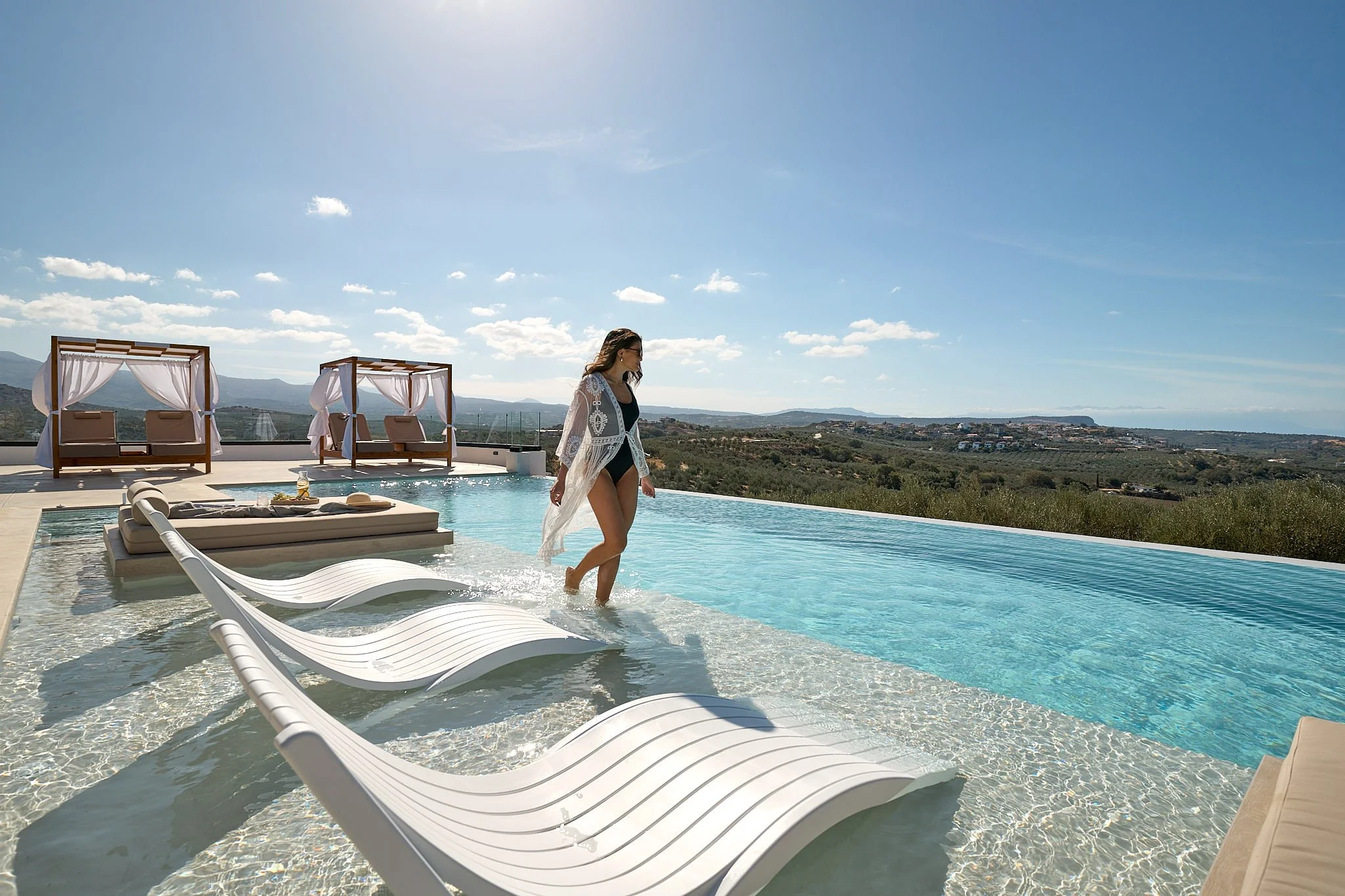 A woman in a swimsuit and sheer cover-up walking in a rooftop infinity pool with lounge chairs and cabanas, overlooking a landscape with distant hills and blue sky.