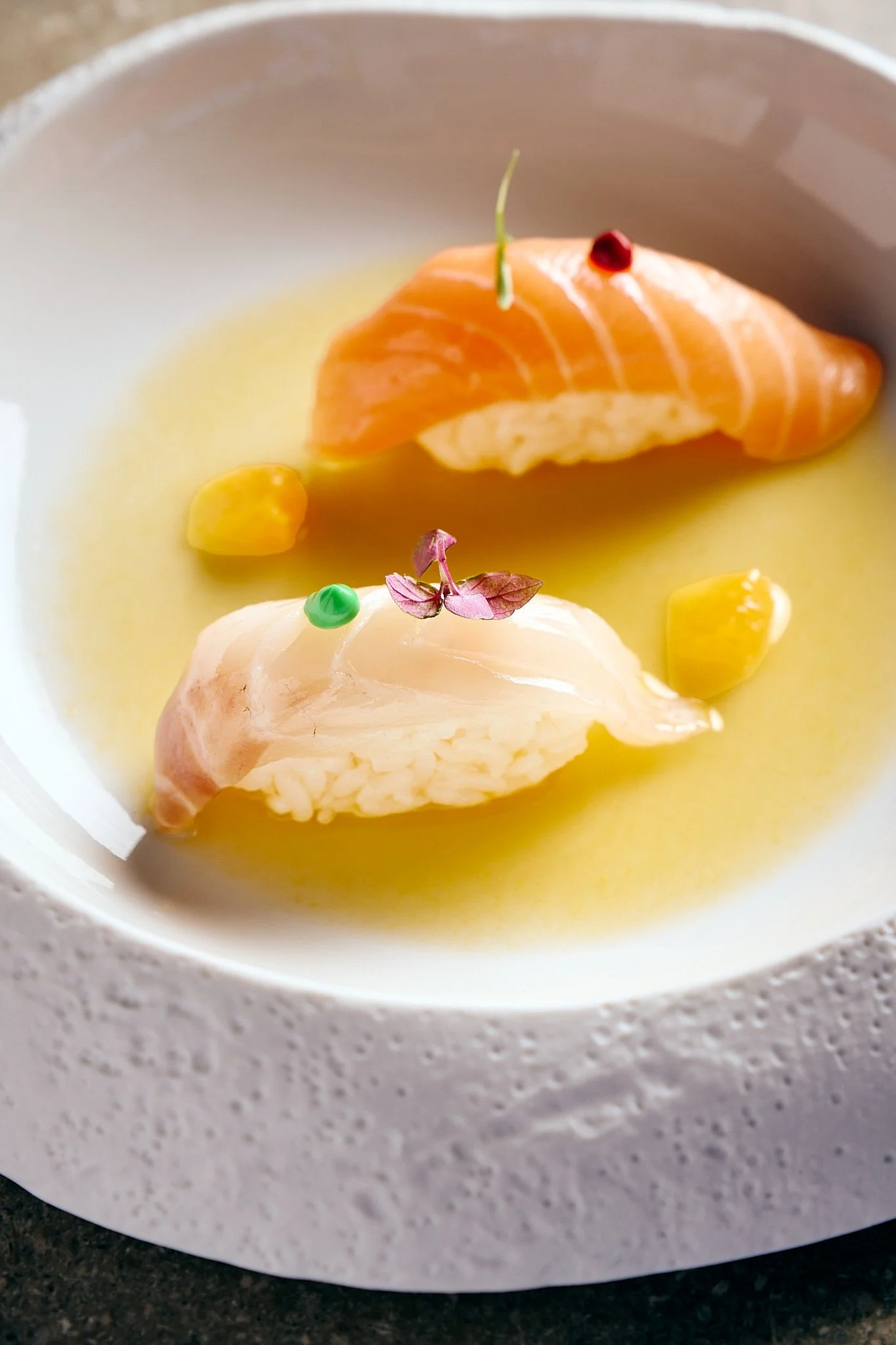 A bowl of yellow broth with two pieces of raw fish, garnished with microgreens and tiny colorful pins, with corn kernels submerged in the broth.