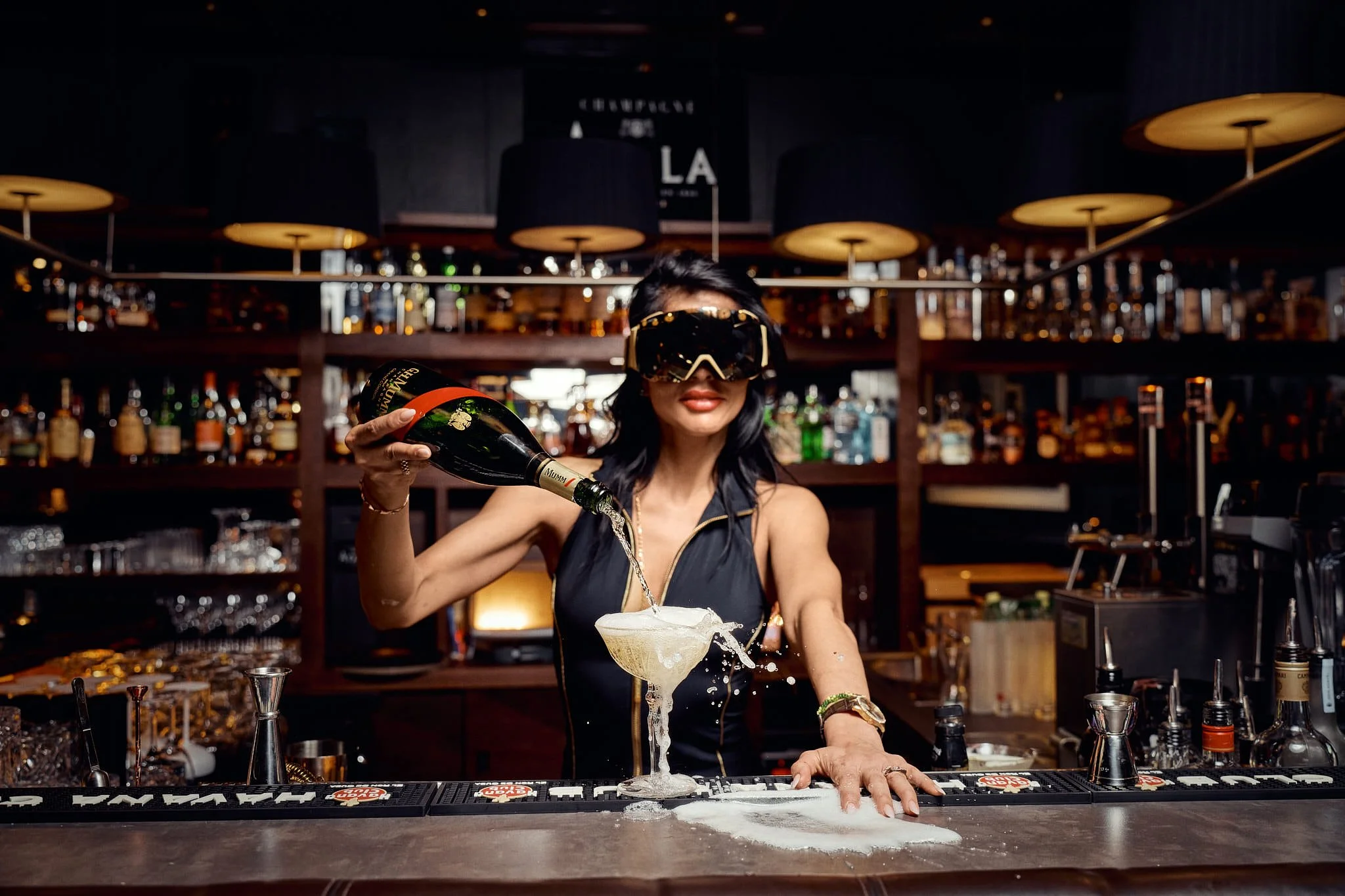 Bartender wearing goggles pouring champagne into a coupe glass with foam overflowing, bar with liquor bottles in background.