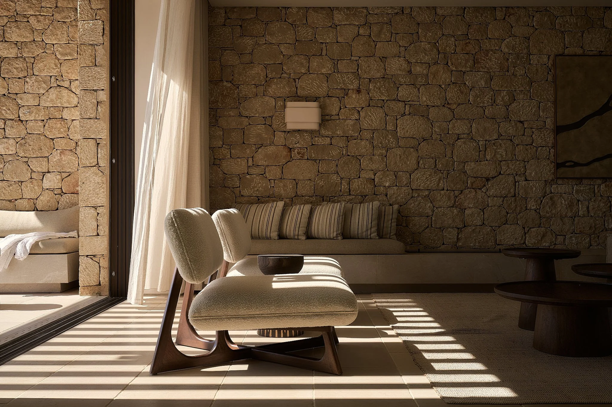 Living room with stone wall, beige armchairs, a couch with striped pillows, and sunlight casting shadows through blinds.