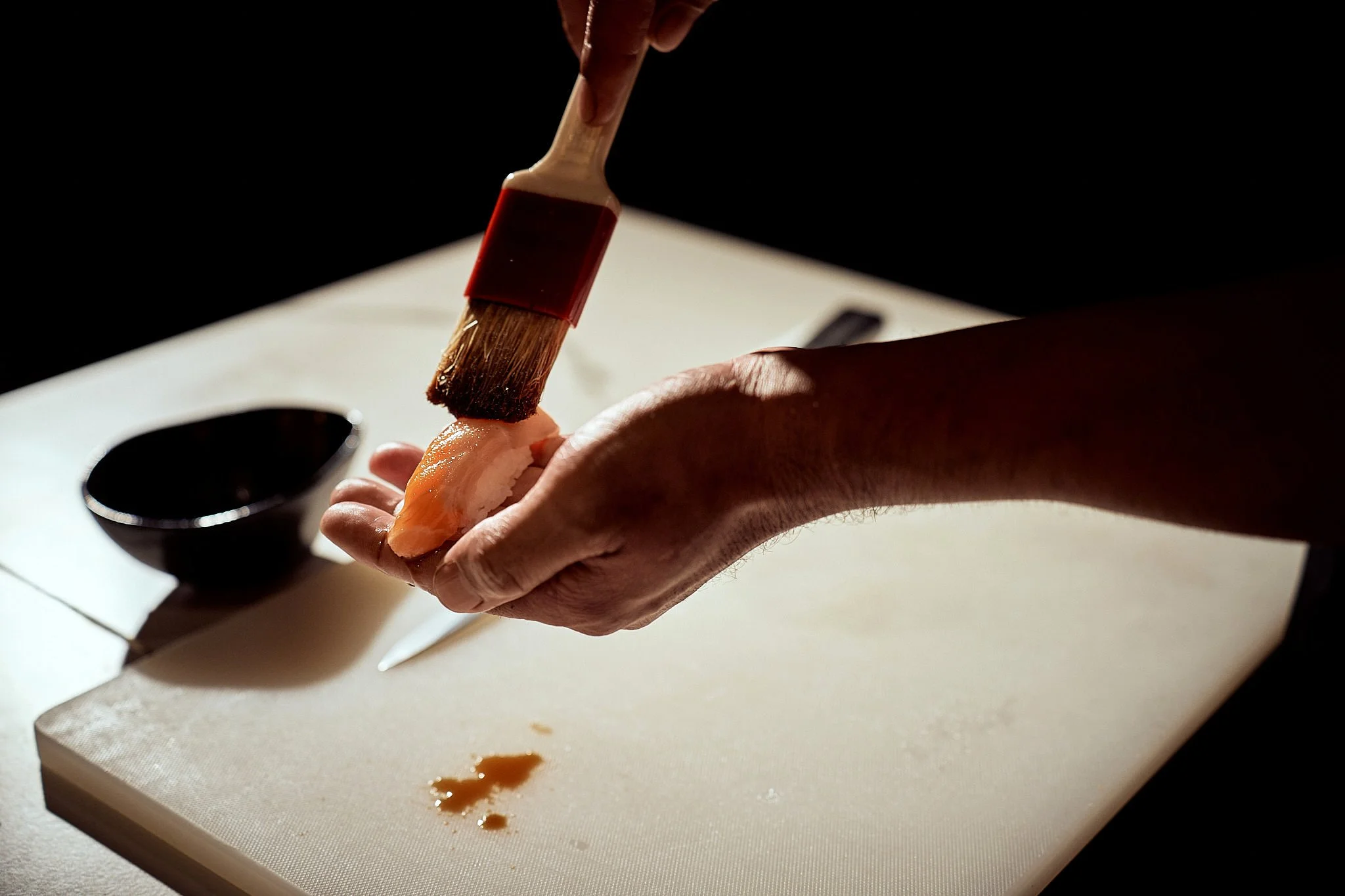 Close-up of a person brushing sauce onto a piece of raw fish on a white cutting board in a dark kitchen environment.