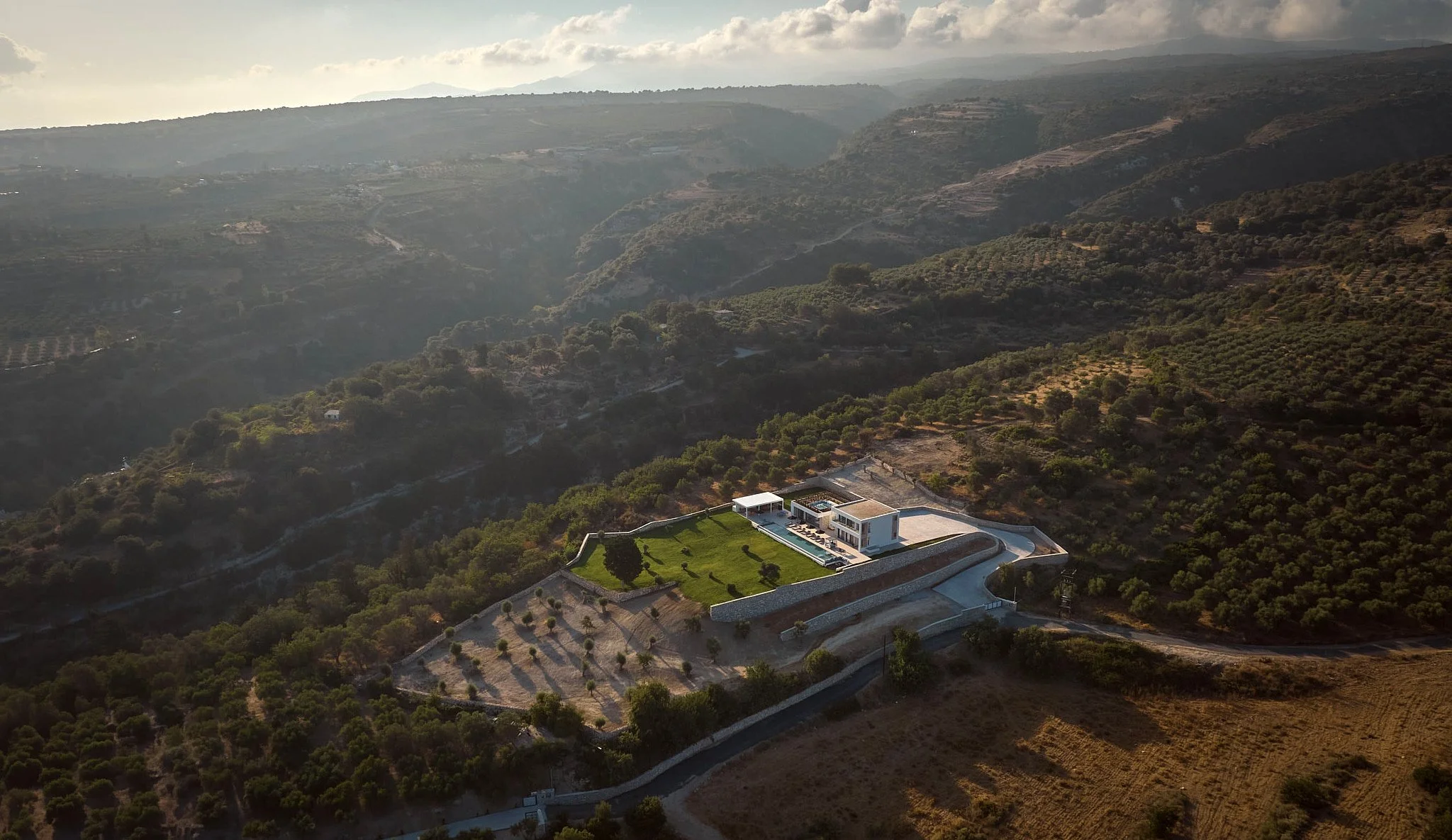 An aerial view of a modern house on a hill surrounded by trees and agricultural land, with rolling hills and mountains in the background under a cloudy sky.