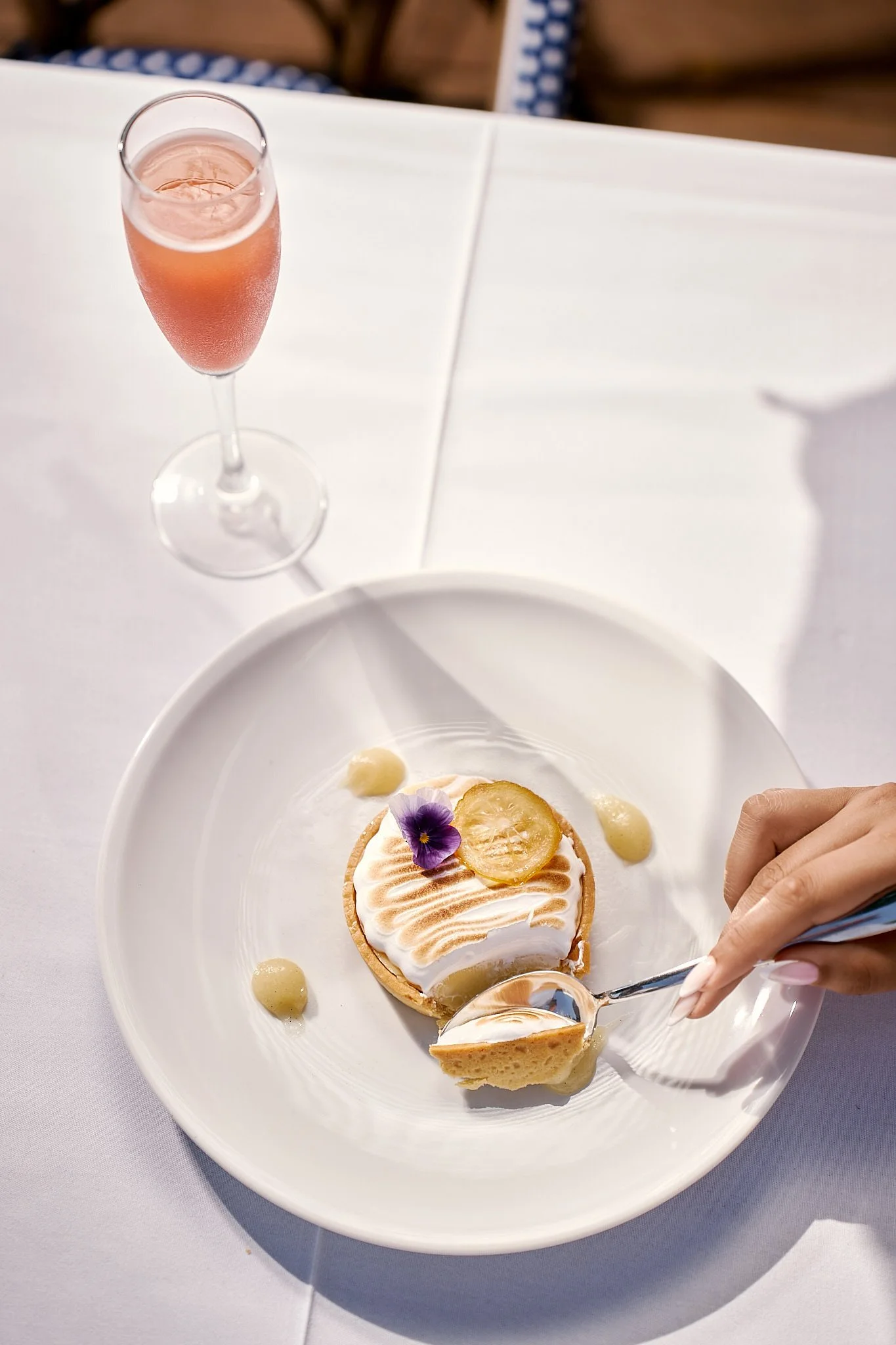 A dessert with a round toasted meringue cake topped with an edible flower and a slice of lemon, garnished with cream and a scoop of lemon sorbet on a white plate. A fork is cutting into the dessert. In the background, a glass of pink sparkling bevera