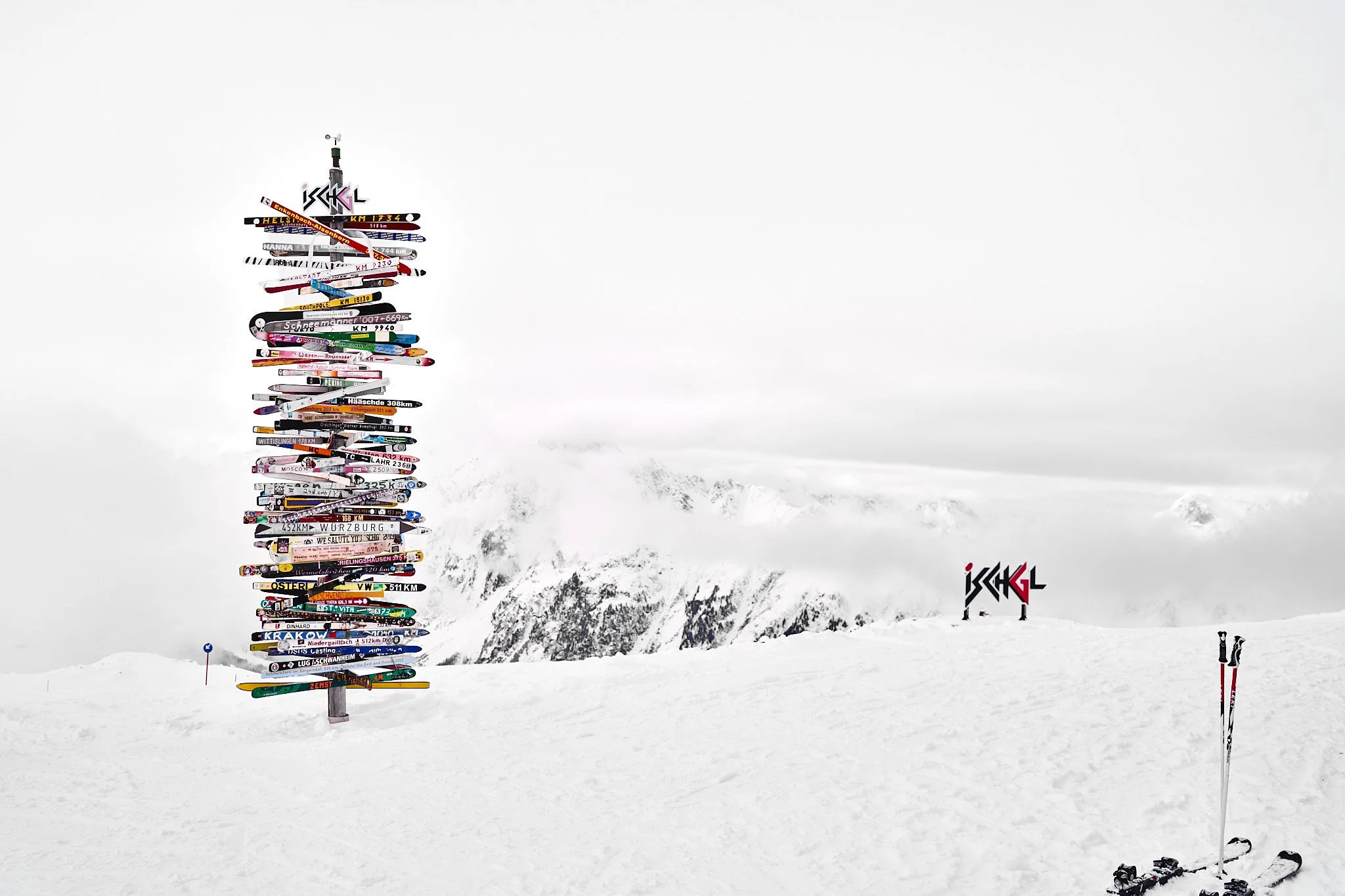 Snow-covered mountain landscape with a tall pole decorated with numerous colorful skis and a sign in the distance with the word 'ISCHGL' on it.