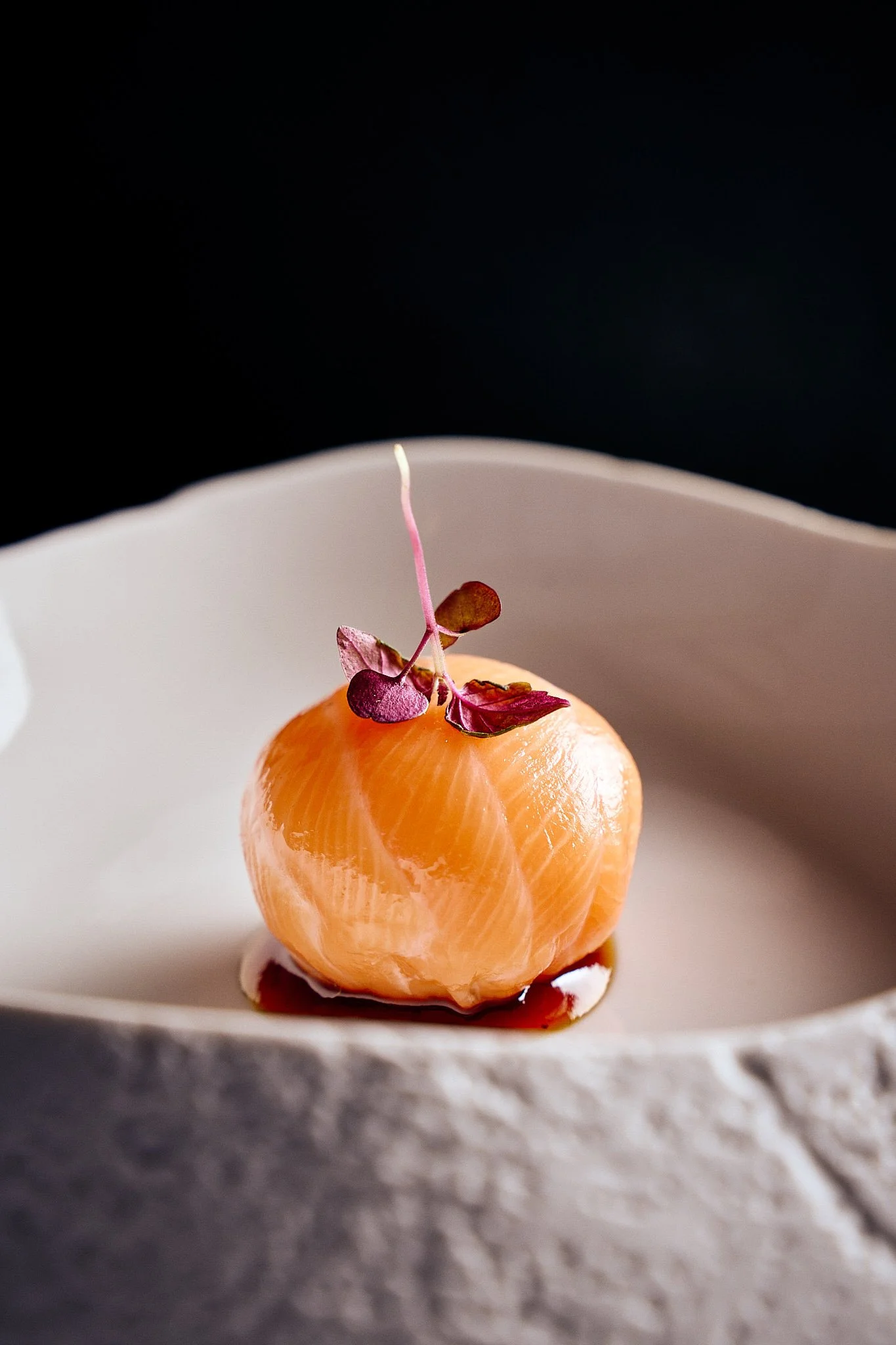 A peeled clementine with a garnish of microgreens on top, placed on a white plate with a dark background.