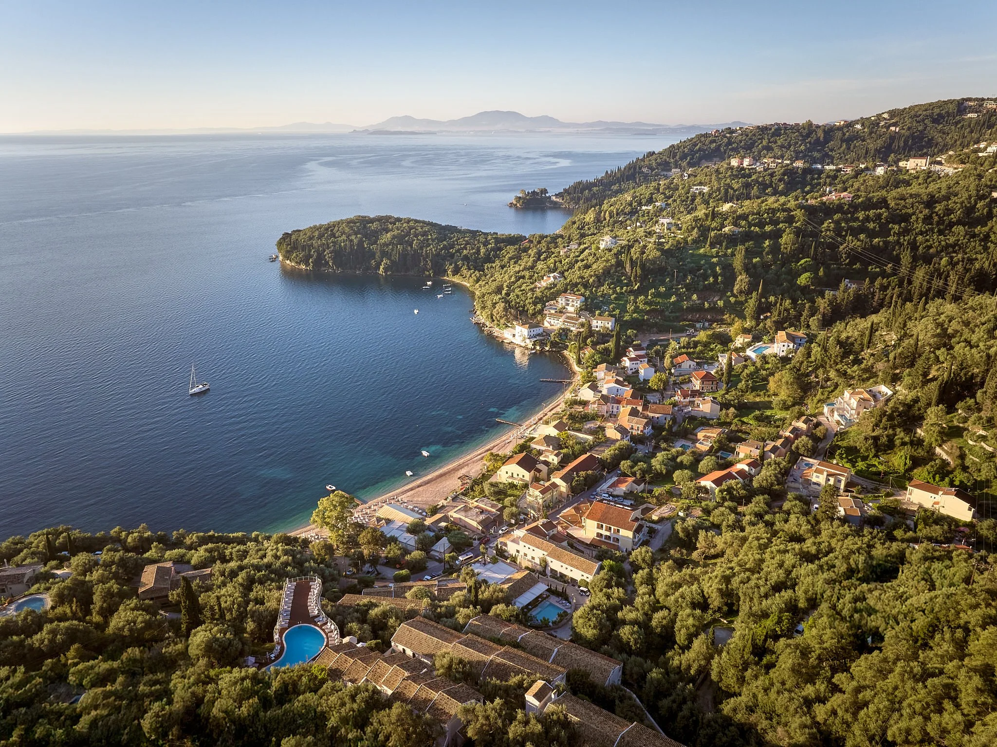 Aerial view of a coastal town with houses and lush greenery along the shoreline and beyond, overlooking a large body of water with a sailboat and several other boats, under a clear sky.