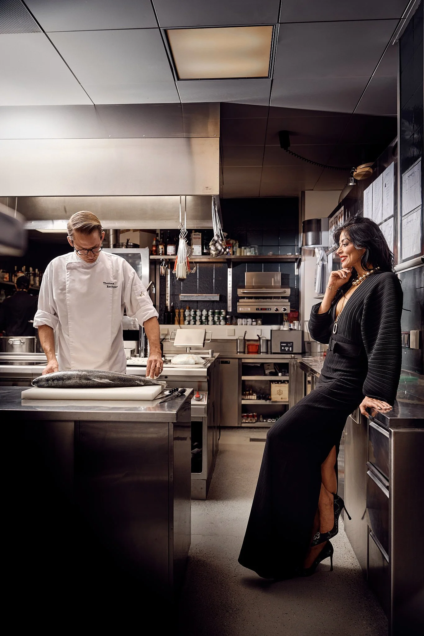 A professional chef in a white coat and glasses working in a restaurant kitchen with a woman in a black dress and high heels sitting on the counter, smiling and looking at the chef.