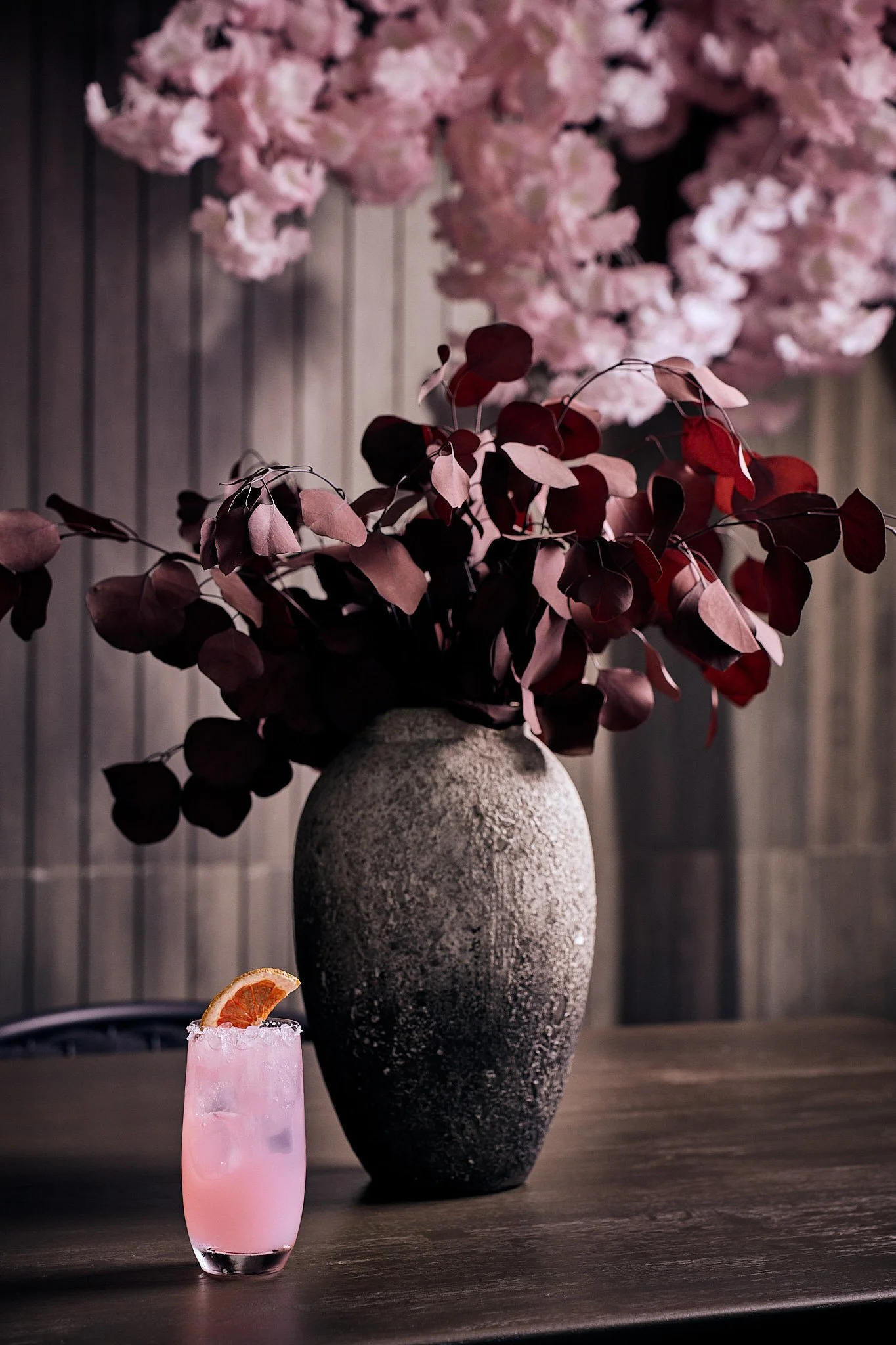 Pink cocktail with a slice of citrus on the rim, placed on a wooden table in front of a large textured dark vase with purple and pink foliage. Blurred pink flowers in the background.