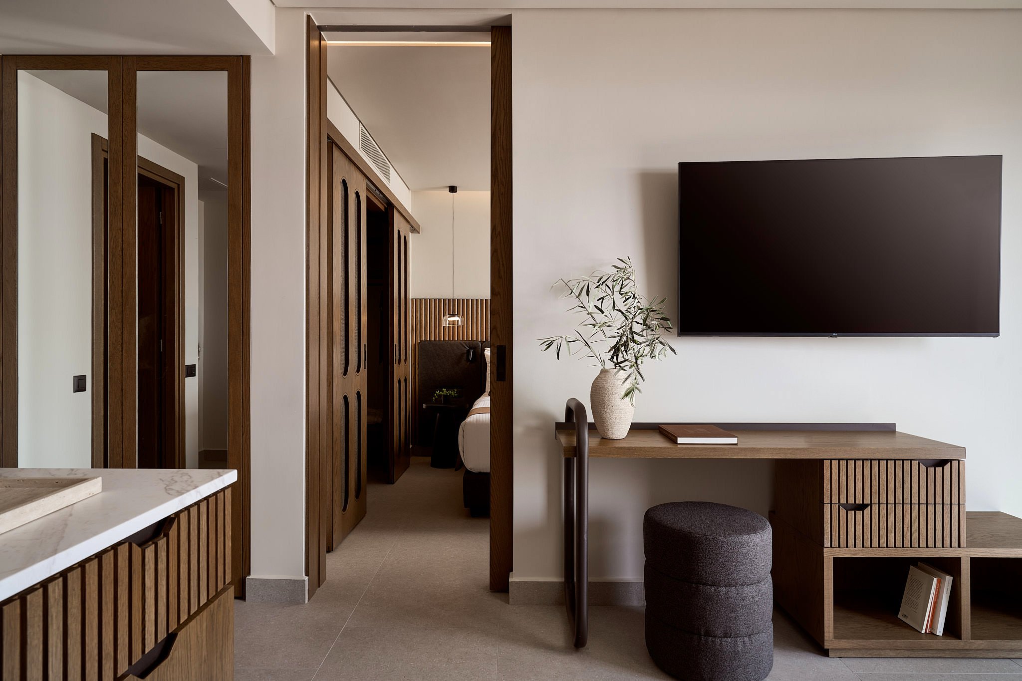 Modern hotel room with a wooden desk, potted plant, large wall-mounted TV, and view of a bedroom with a headboard and pendant light.