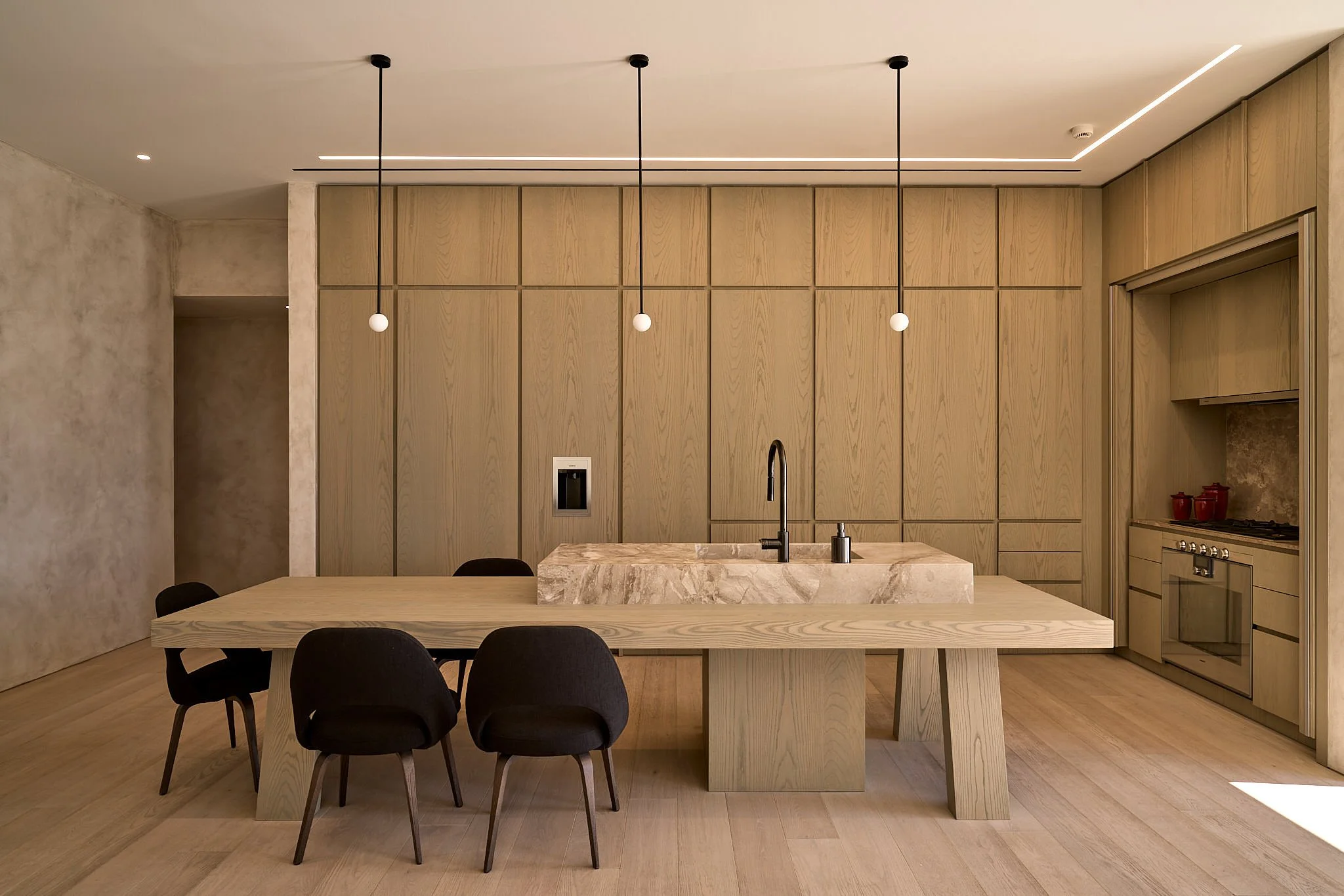 Modern minimalist kitchen with wooden cabinetry, a marble kitchen island with a built-in sink, black pendant lights hanging above, and black chairs around a wooden dining table.