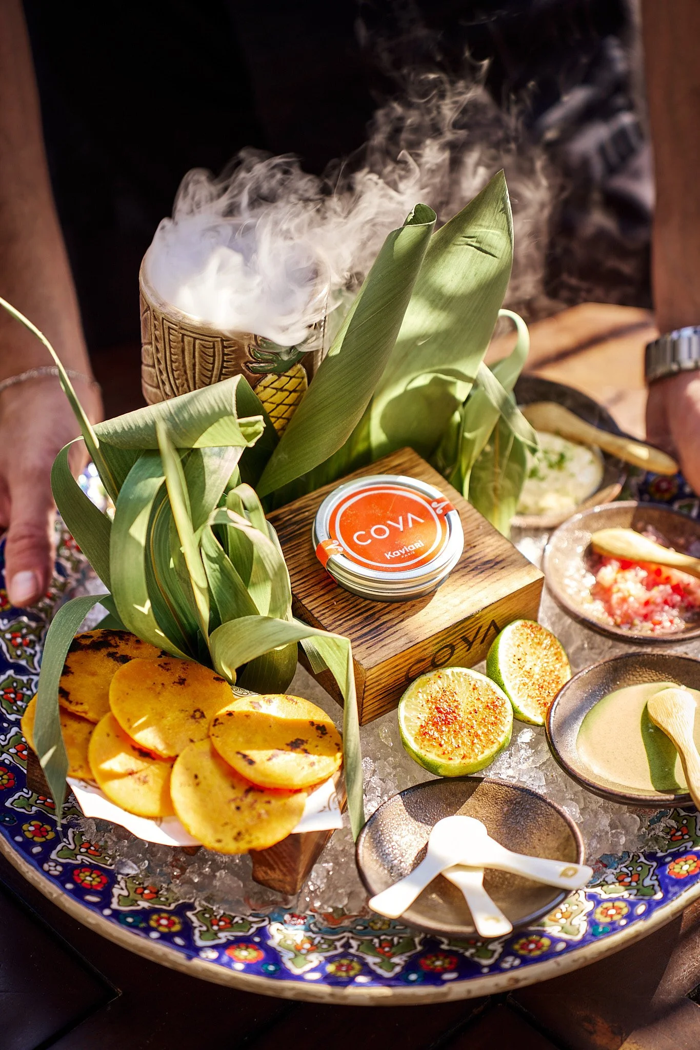 A traditional Mexican molcajete with various salsas, grilled pineapple slices, limes, and a steaming cup of hot beverage surrounded by foliage on a decorative plate.