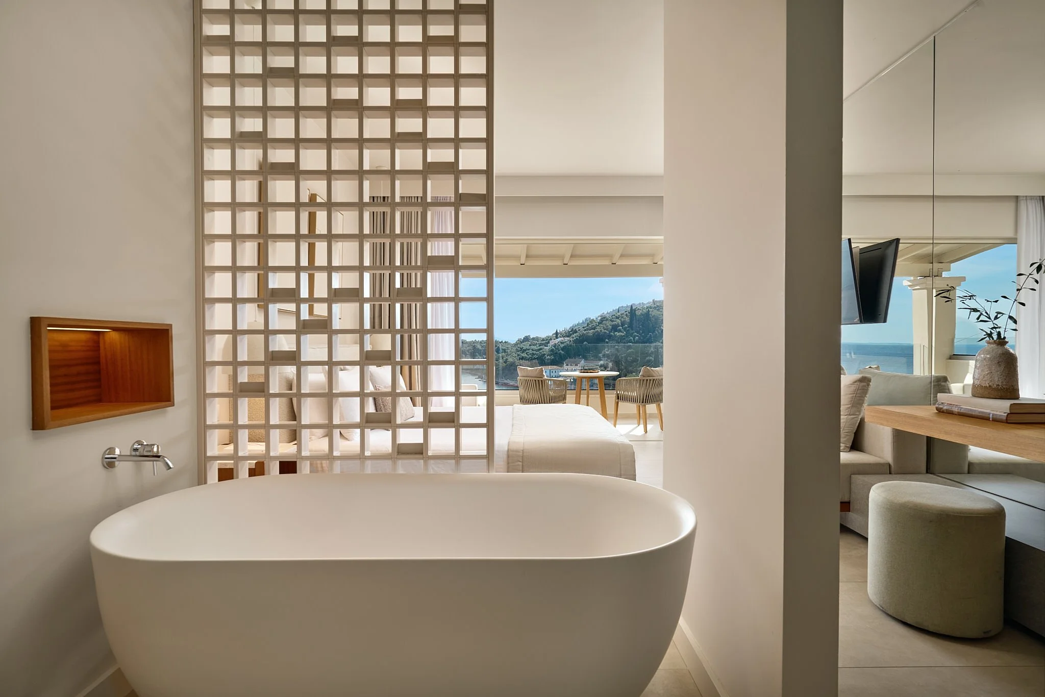 Modern hotel room with a freestanding bathtub, sitting area with views of the ocean and hills, and balcony with outdoor seating
