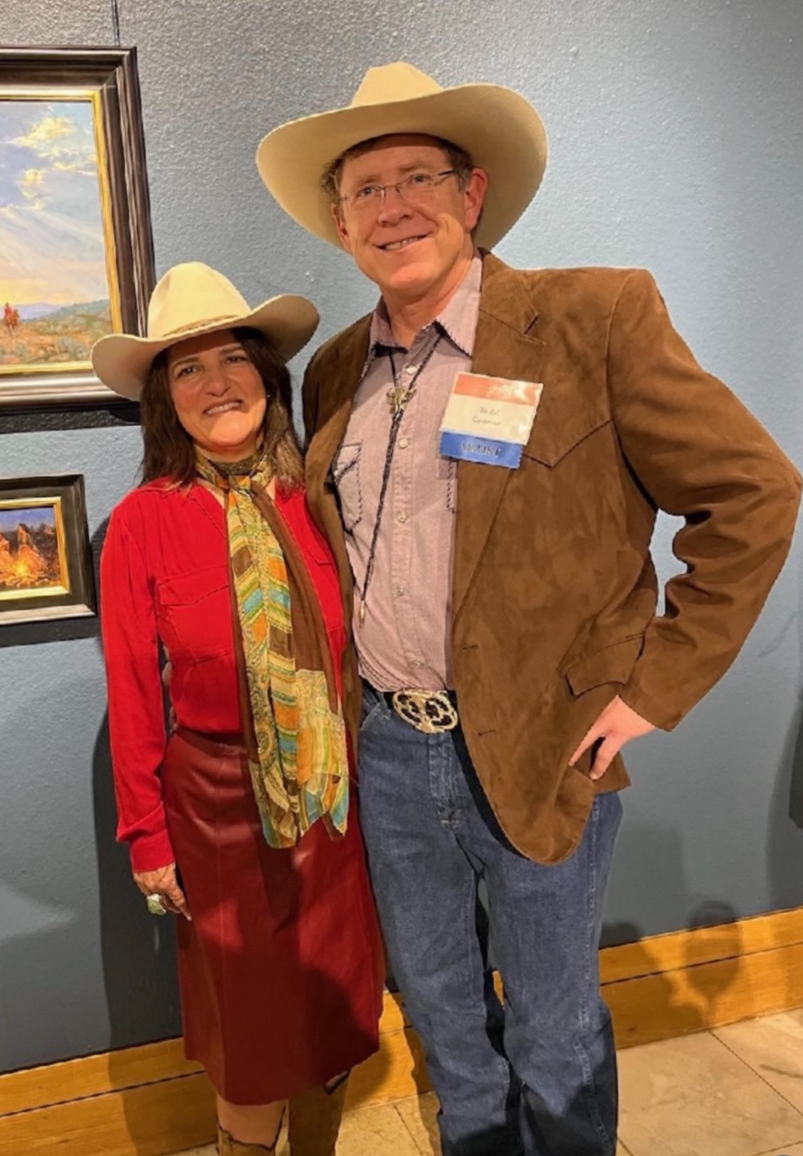 A man and woman dressed in Western attire, both wearing cowboy hats, standing together in front of framed landscape paintings on a blue wall.