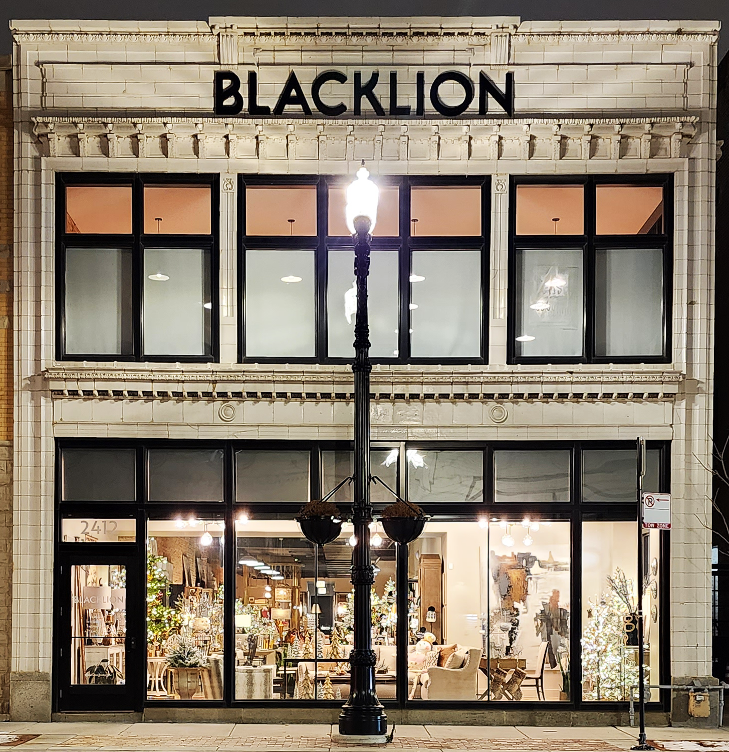 Storefront with large glass windows displaying home decor and Christmas decorations, illuminated at night, with a black street lamp in the foreground and the name 'BLACKLION' on the building's upper facade.