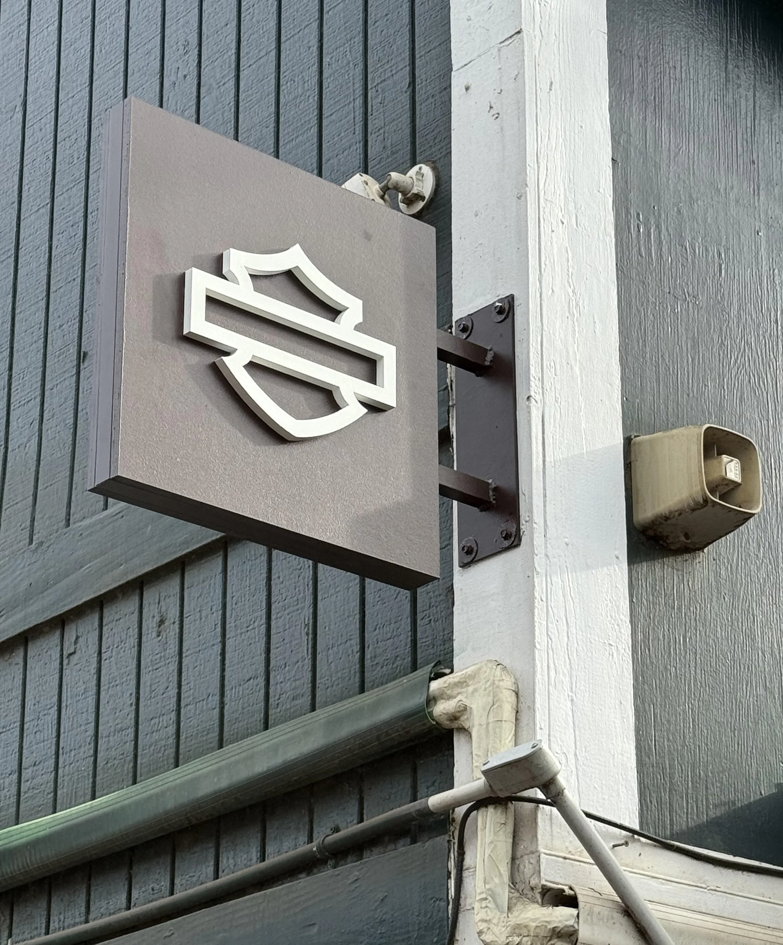 Close-up of a building exterior featuring a mounted Harley-Davidson logo sign, part of the wall, a small surveillance camera, and a beige utility box.
