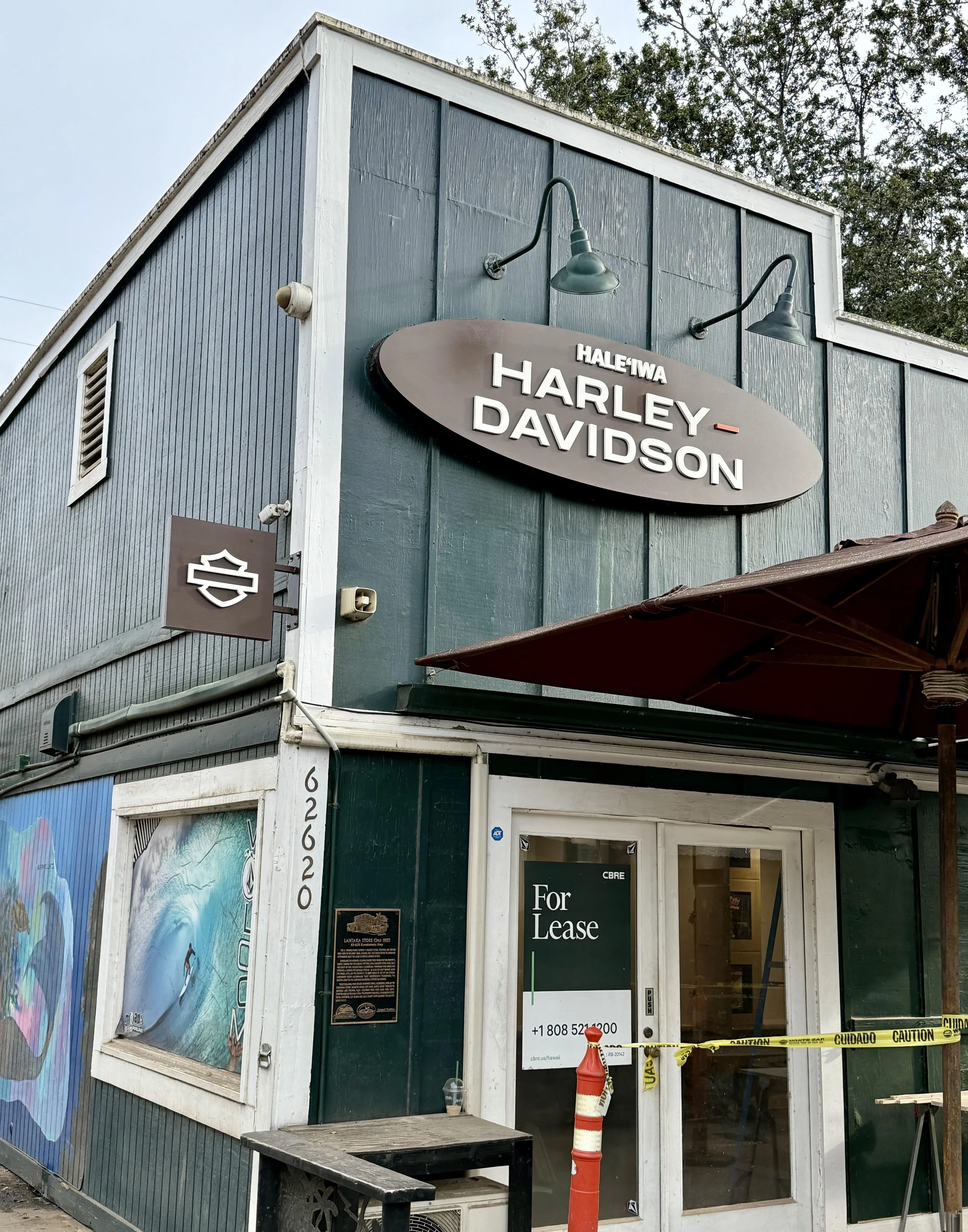 Two-story commercial building with a teal and white exterior, featuring Harley Davidson signage and a 'For Lease' sign on the door, partially obstructed by caution tape and orange traffic cone.