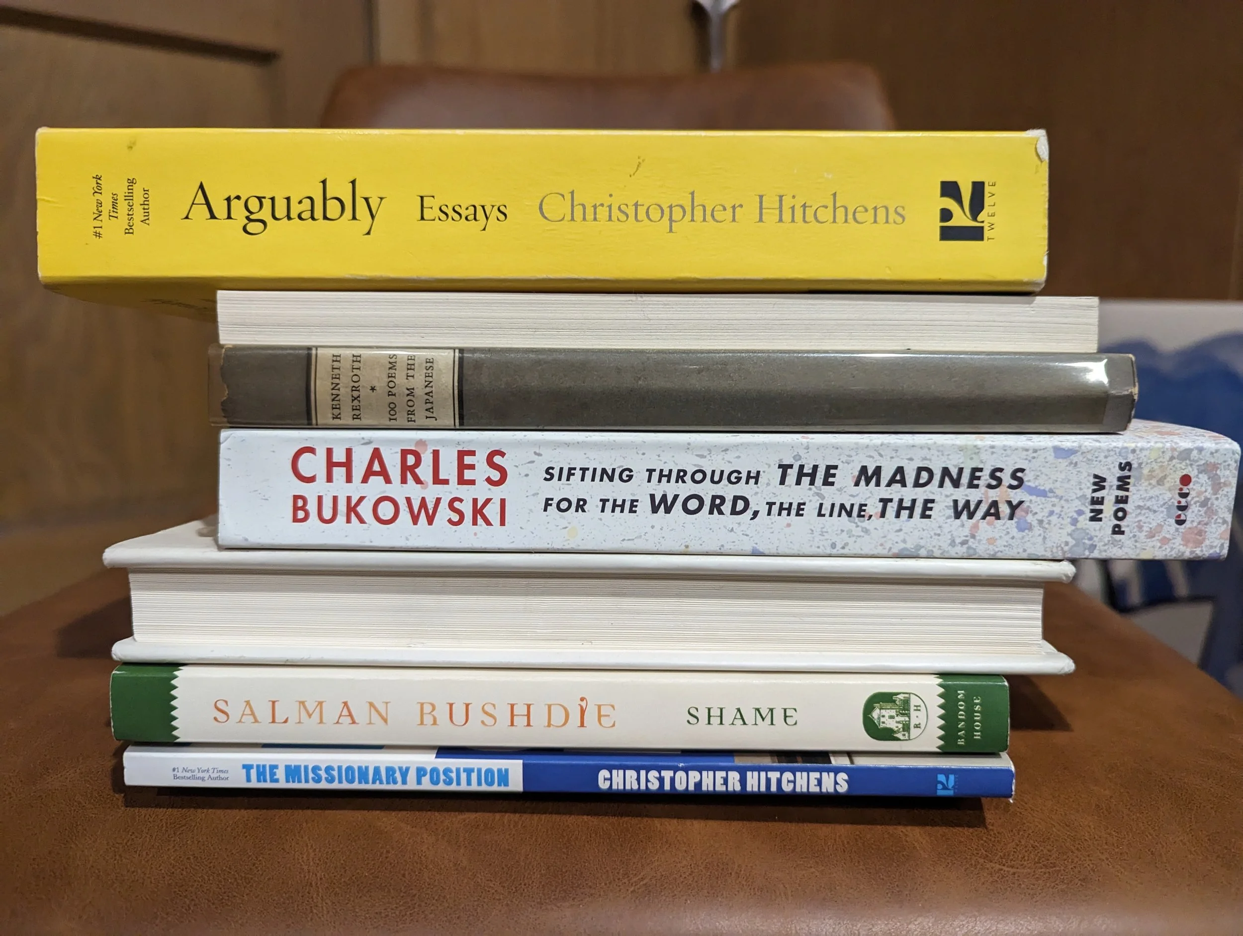 Stack of eight books on a table, with titles including "Arguably Essays" by Christopher Hitchens and "Shame" by Salman Rushdie.