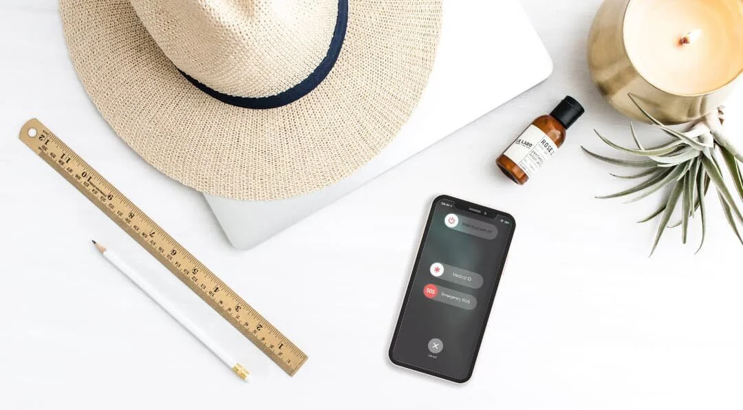A minimal travel-work flatlay with a sun hat, iPhone, laptop and calming essentials — a soft, airy scene for pausing your business rhythm.