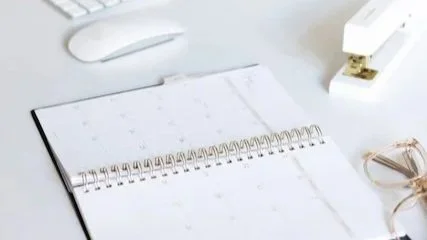 A minimal white desk with an open planner, soft natural light and calm workspace details — a gentle moment of clarity and reset.