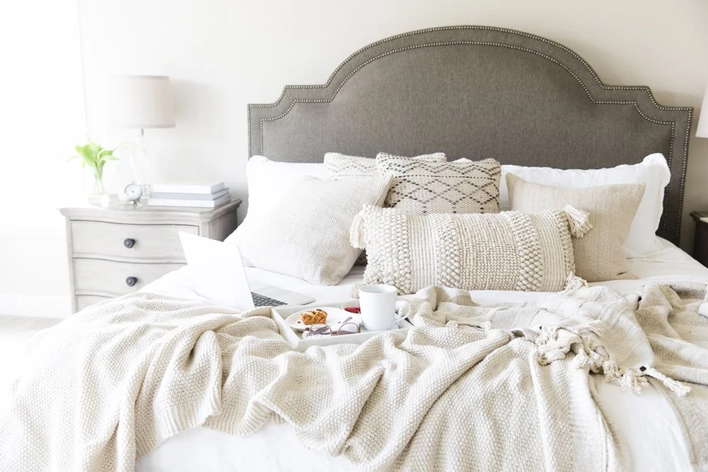 Neutral bedroom interior with layered cream bedding, textured cushions and soft morning light, creating a calm, restful atmosphere.