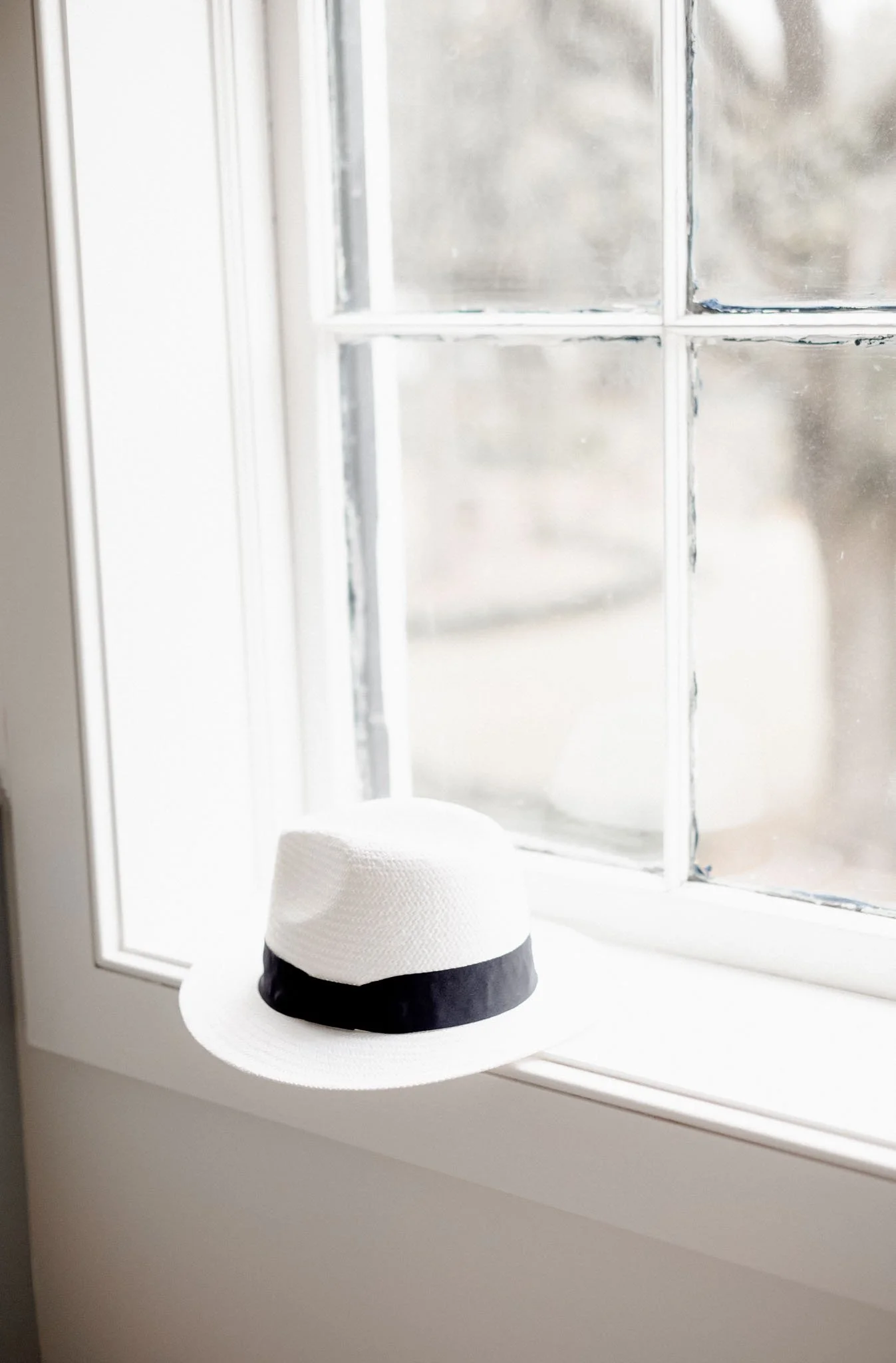 White window sill with a crisp white Panama hat and softened exterior greenery beyond the glass, simple and calming design detail.